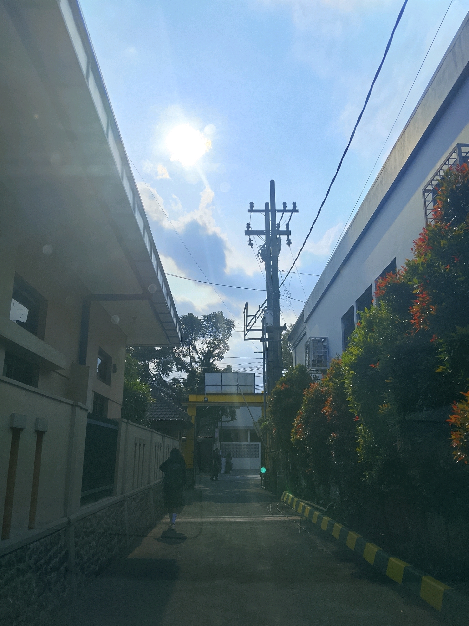 The image depicts a street scene in Malang, Indonesia, under a bright, sunny sky with scattered clouds. On the left, a light-colored building with a modern, angled roofline extends into the frame, casting a shadow. A stone wall with vertical elements runs along the side of this building. On the right, a white building with windows and an air conditioning unit is visible, partially obscured by lush green trees with red-tinged leaves. A power pole with electrical wires stands prominently in the center of the street. The road is paved and marked with yellow and black safety barriers along the right side. In the foreground, a person wearing dark clothing and a backpack walks away from the camera down the street. Further down the street, another person can be seen, and beyond them, a yellow archway is visible. The overall impression is a daytime view of a street in a built-up area, possibly a residential or institutional complex.
