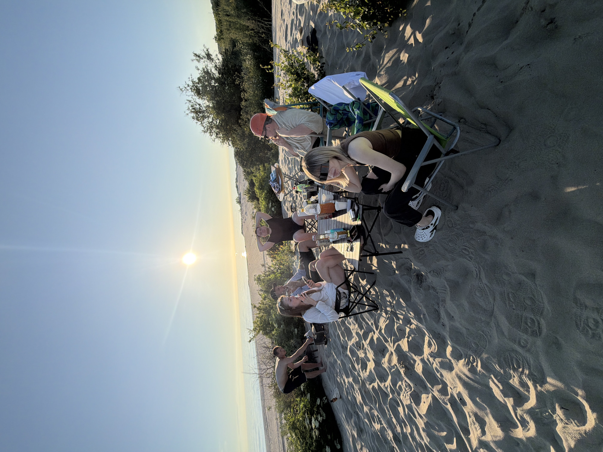 A group of six people are gathered on a sandy beach near the sea, likely in Povorovo, Russia, during sunset. The sun is low in the sky, casting a warm glow and long shadows across the sand. The group appears to be enjoying a casual outdoor gathering, with some seated in camping chairs around a low table. On the table, there are bottles of drinks, cups, and what looks like food. One person is tending to a small grill or fire pit. The environment is natural, with sand dunes, sparse vegetation, and the sea stretching to the horizon. The overall mood is relaxed and social.
