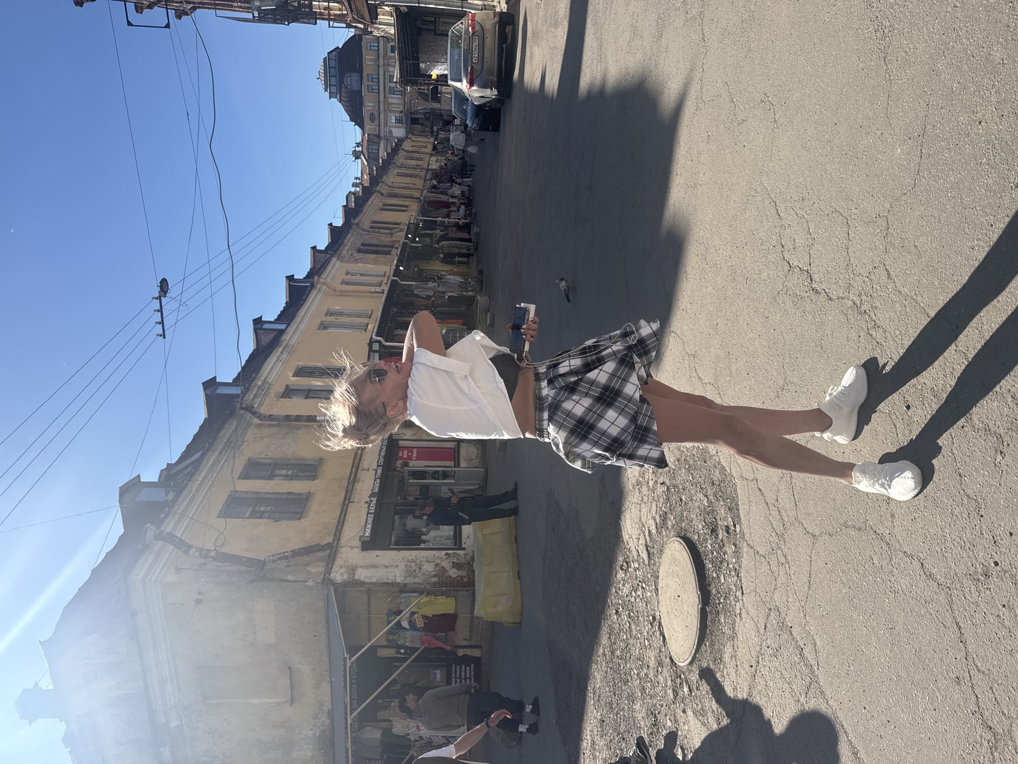 A blonde woman in a white top and plaid skirt is captured in a candid moment, her arms raised as if dancing or stretching. She is holding a phone in her right hand. The scene is an outdoor marketplace with buildings lining a street. The architecture suggests an older European city. Sunlight casts long shadows, indicating it's likely late afternoon. The weather appears to be clear and sunny. The woman's expression is joyful and energetic.
The street is paved and shows signs of wear, with a manhole cover visible on the right side. In the background, stalls with merchandise are set up along the buildings, suggesting a bustling commercial area. Several people are milling about, some appearing to be vendors or shoppers. A car is parked further down the street. Overhead, power lines crisscross against a clear blue sky.
Visible text on the buildings includes Cyrillic script, consistent with a Russian location. One sign reads "НИЖНЕЕ БЕЛЬЕ" (lingerie) and another appears to advertise "СПОРТИВНАЯ ОДЕЖДА" (sportswear). The overall atmosphere is lively and casual, with the woman's movement adding a dynamic element to the otherwise static scene of street commerce. The sunlight glinting off the buildings and the shadows on the ground contribute to a bright, warm ambiance.