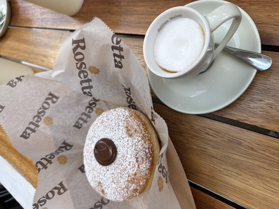 A round pastry, dusted with powdered sugar and topped with a dollop of dark brown chocolate filling, rests on white parchment paper printed with the "Rosetta" logo and emblem. To its right, a white ceramic cup containing a foamy beverage, likely coffee, sits on a matching saucer with a small silver spoon. The inside rim of the cup is marked "Julius Meinl". All items are positioned on a light brown wooden slatted table. The background is blurred. The scene depicts a breakfast or snack setting at a Rosetta Bakery establishment in Miami, Florida, United States.