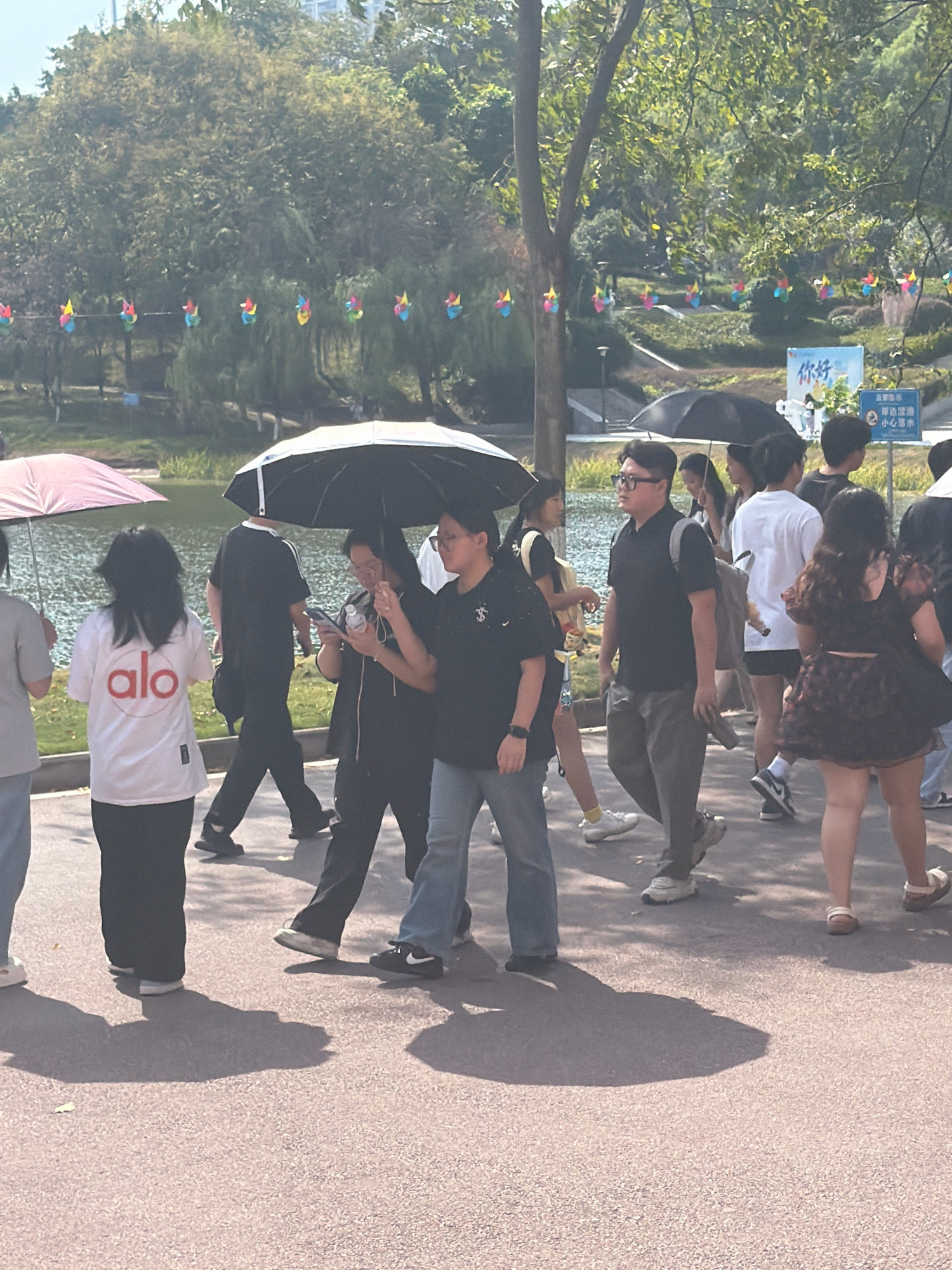 In Jiangjin, China, a group of people are walking along a paved path beside a body of water. A few individuals are holding umbrellas. In the background, trees line the water's edge, and a row of colorful pinwheels are strung up, possibly as decoration. There is a sign in the background with Chinese characters. The scene appears to be a park or recreational area.