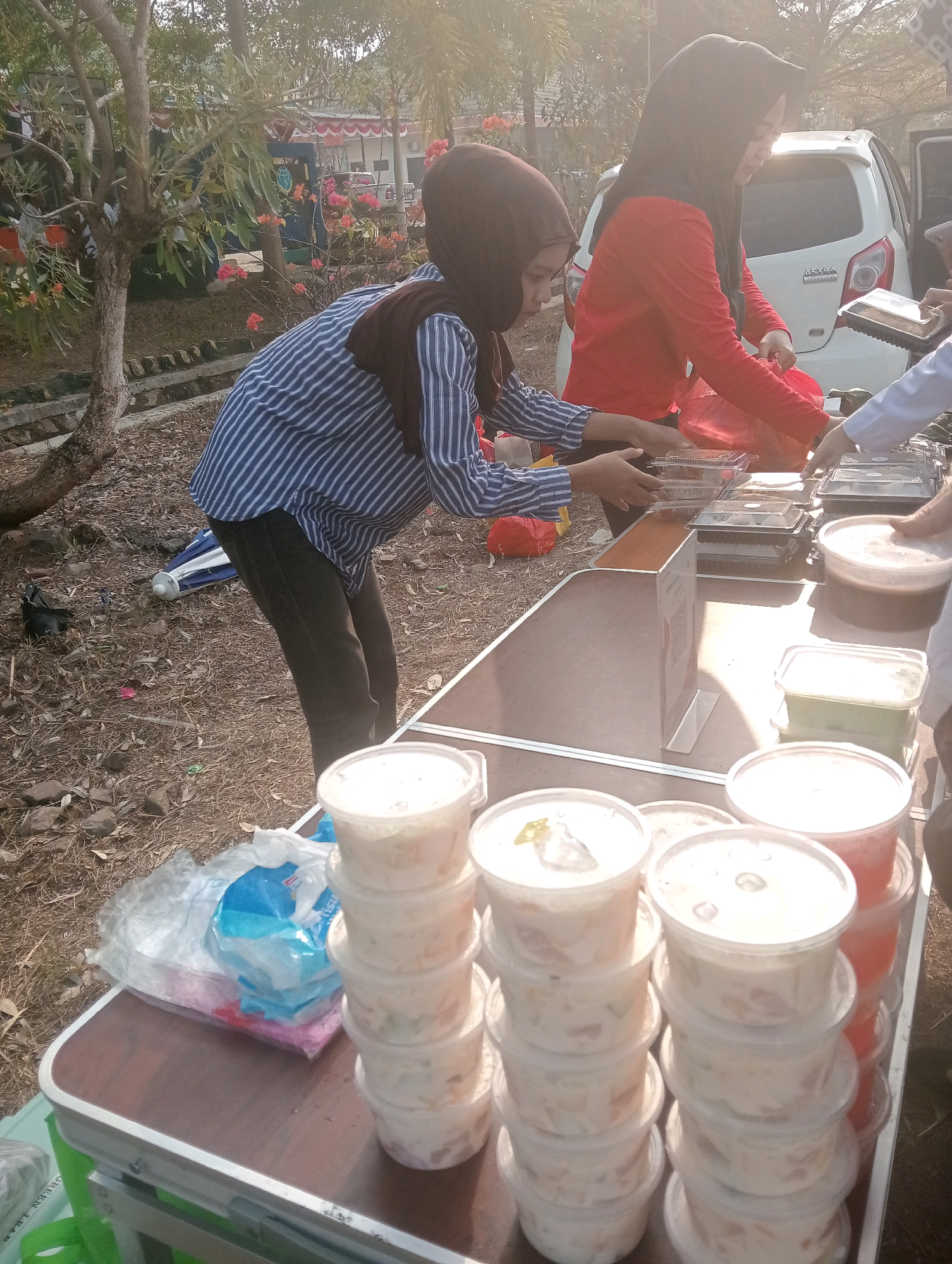 Two veiled women are standing behind a folding table, appearing to be selling food, in what seems to be an outdoor event or market in Sidoharjo, Indonesia. The woman on the left wears a brown hijab and a blue and white striped long-sleeved shirt. She is reaching for a clear plastic container of food. The woman on the right wears a red long-sleeved shirt and a black hijab. She is handing a red plastic bag to someone off-camera. The table is laden with stacked clear plastic containers filled with food, some of which appear to be desserts or salads. There are also some opaque plastic containers. In the background, a white car with "ASTRA TOYOTA" branding is visible. The ground is covered with dry leaves and dirt, and there are trees and some flowering plants to the left of the scene. The lighting suggests it is daytime, possibly late afternoon given the warm light.