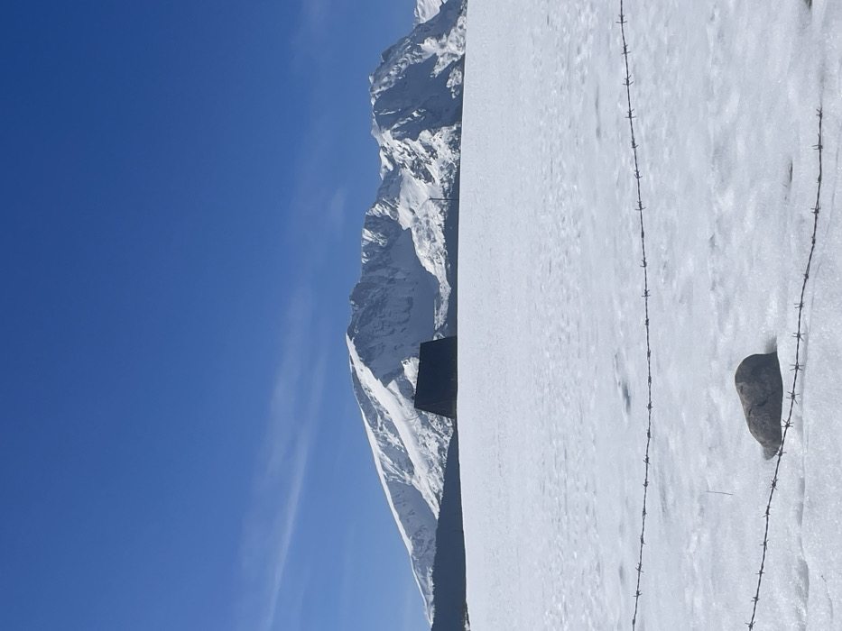The image depicts a vast, snow-covered winter landscape under a clear blue sky, situated in Palež, Montenegro. In the immediate foreground, two parallel strands of rusted barbed wire stretch horizontally across the lower portion of the frame, partially obscured by snow. A single, dark gray rock, approximately football-sized, rests on the snow just above the lower barbed wire strand, slightly to the left of the frame's center. The midground is dominated by an expansive, uninterrupted field of white snow. Centered horizontally in the distance, a small, dark-colored building with a steeply pitched, A-frame or triangular roof stands out against the snow, appearing solitary. Beyond the building, the background features a long range of rugged, snow-capped mountains, exhibiting visible rock formations and deep snowdrifts. The sky above is uniformly bright blue with only a few faint, wispy cirrus clouds visible in the upper left quadrant. No individuals or overt actions are present in the scene, which conveys a sense of stillness and cold.