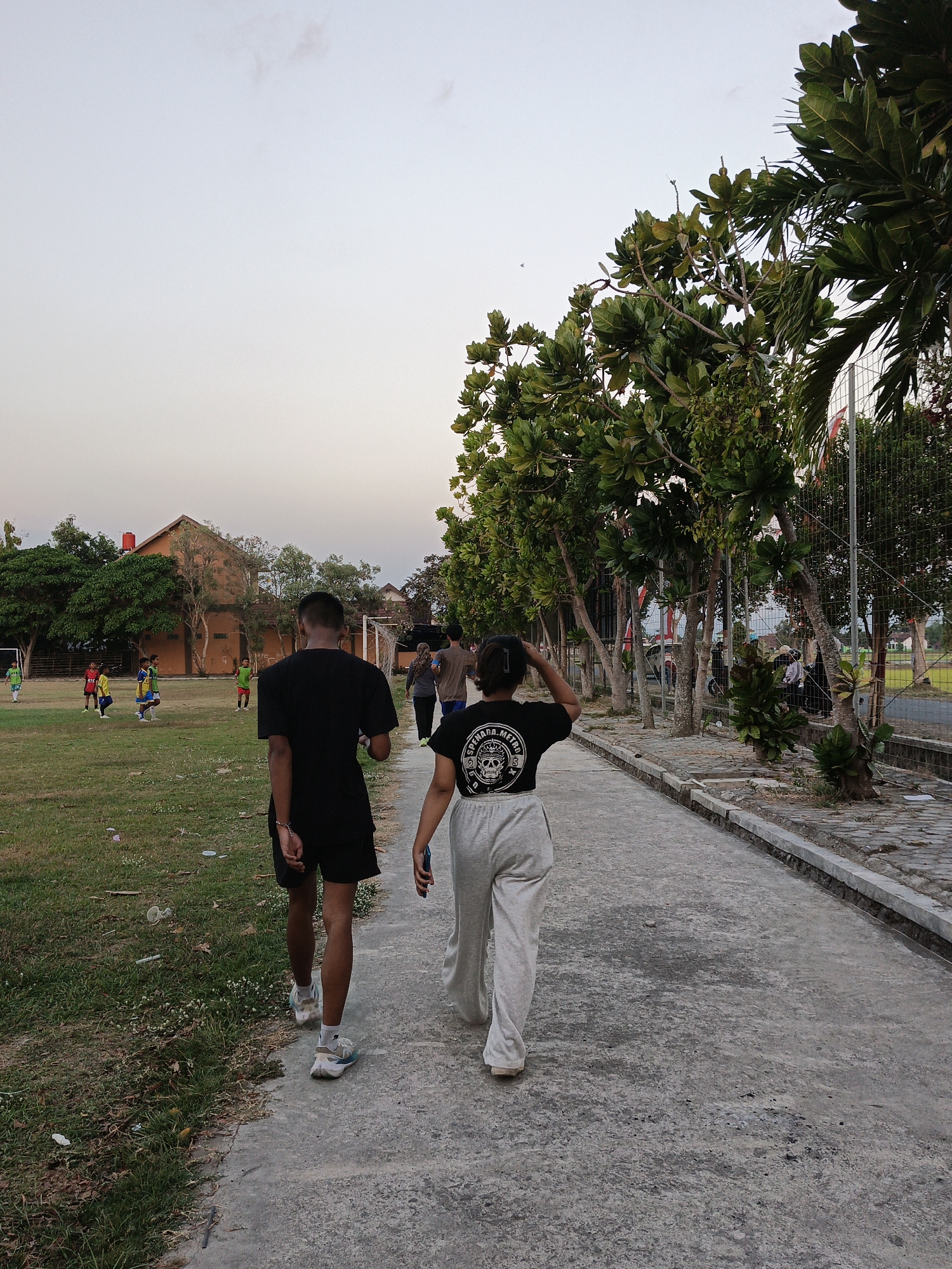 Two individuals, one male and one female, walk away from the camera on a paved path adjacent to a grassy field. The male, wearing a black t-shirt and black shorts, walks slightly ahead and to the left. The female, wearing a black t-shirt with a graphic design and light gray sweatpants, walks to the right. In the background, children are playing soccer on a field in front of a two-story building. Trees and foliage line the path and the field. The sky is a pale blue with a few wispy clouds. The scene appears to be a public park or recreational area in Planggu, Indonesia.