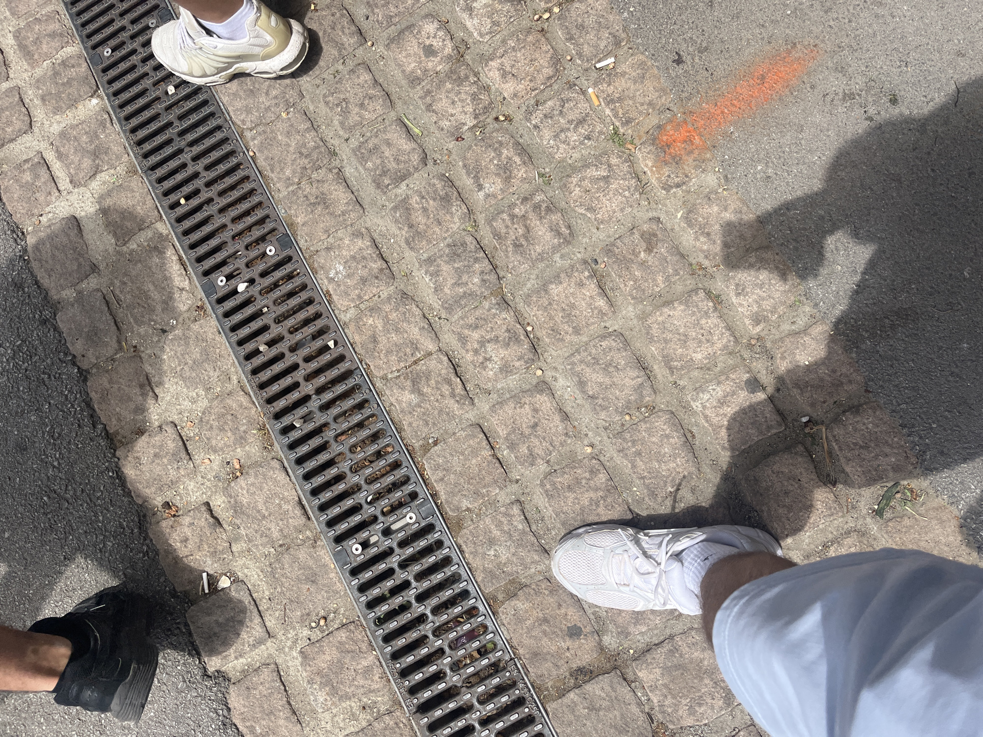 The image captures a ground-level view of a European-style urban environment, likely in Blagoevgrad, Bulgaria, based on the cobblestone pavement. A metal drainage grate runs diagonally across the frame, filled with debris like cigarette butts and leaves. Two individuals are visible, each represented by a foot and shoe. One person is wearing a white sneaker with a sock visible, paired with light-colored shorts. The other individual wears a black shoe and a dark-colored sock. An orange marker on the asphalt indicates some form of marking or designation. Shadow indicates that a third individual took the picture.