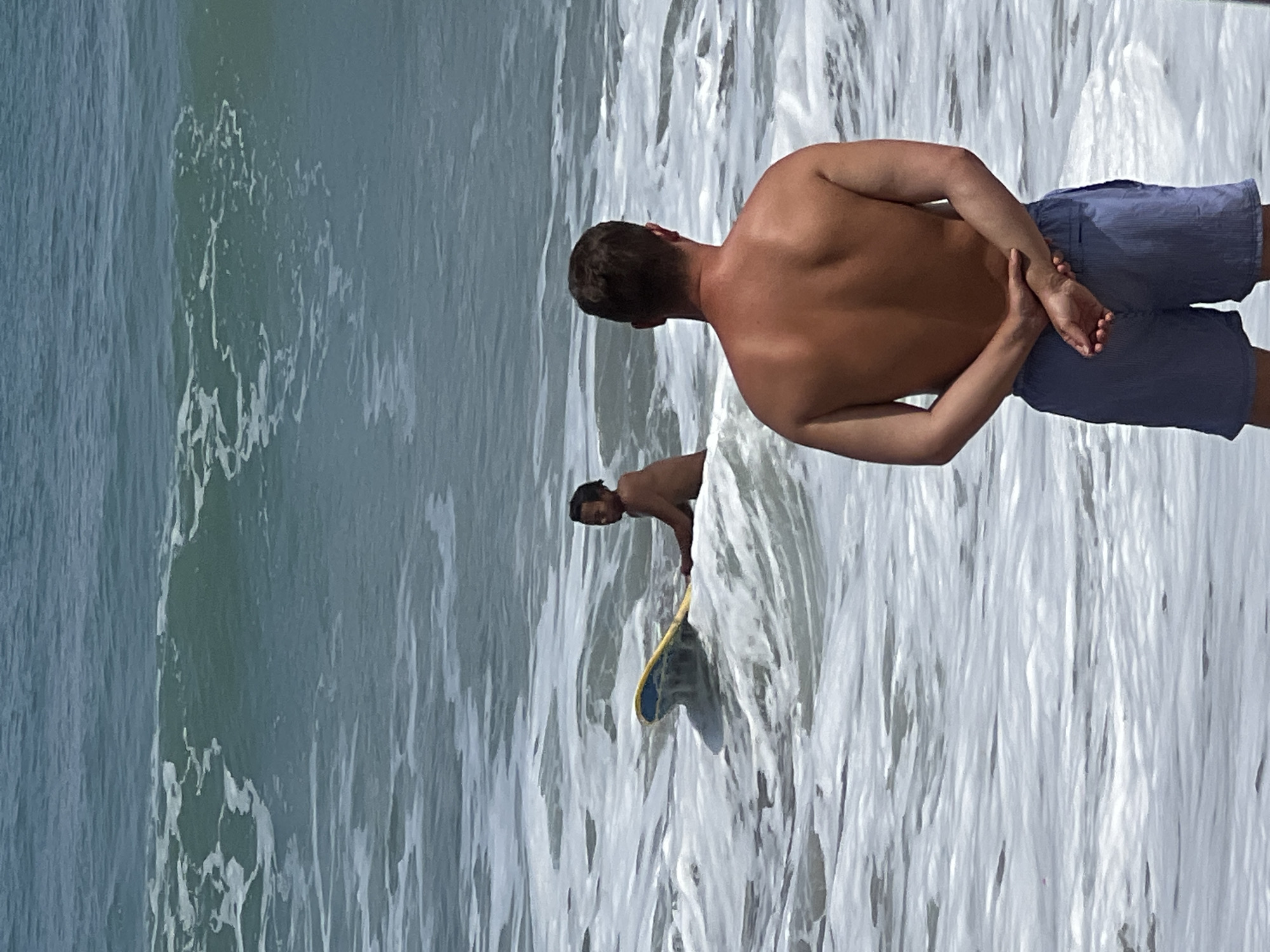 In the ocean off Kuta, Indonesia, two men are present. The man in the foreground is facing away from the camera, standing in the shallow water at the shoreline, with his hands clasped behind his back. He is wearing blue striped swim shorts. In the background, a second man is in deeper water, sitting on a yellow and blue surfboard. He is looking to his left, away from the camera. The ocean has moderate waves with white foam.