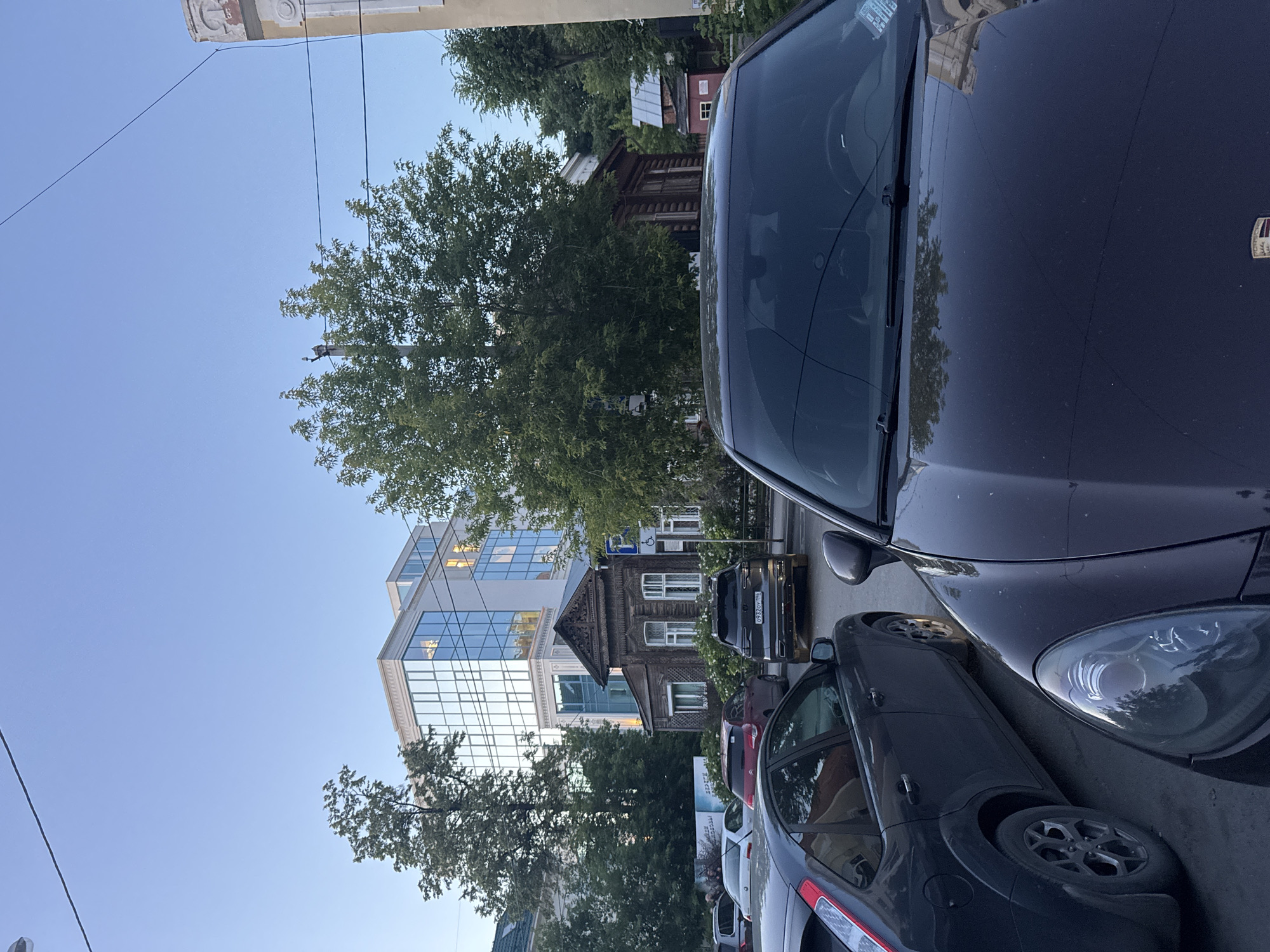 The image shows a street scene in Yekaterinburg, Russia, likely in an urban residential or mixed-use area. The foreground is dominated by the hood and windshield of a dark-colored Porsche Cayenne, with its reflection of trees and sky visible on the hood. To the left of the Porsche is a dark grey Ford Focus. Behind these vehicles, a dark wooden traditional Russian house with white trim is situated. Behind the wooden house, a modern, multi-story building with a glass facade is visible. Trees are present throughout the scene, some mature and large, others smaller and more shrub-like. The sky is clear and light blue, suggesting it is either early morning or late evening. A license plate on a black SUV parked behind the wooden house reads "0932OV 196". A blue and white sign with text is also visible on a pole near the SUV. The overall impression is a street with a mix of old and new architecture.