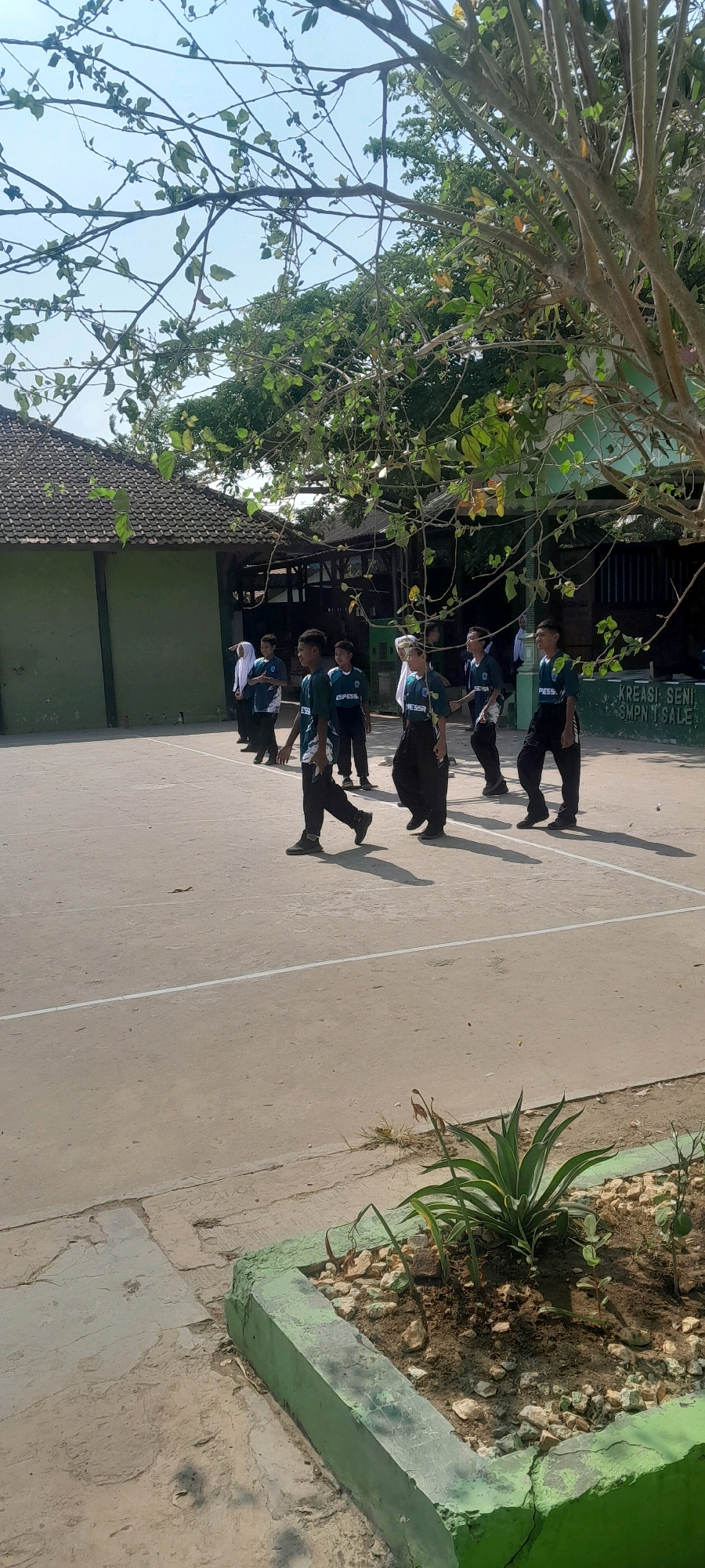 The image shows a group of students, likely middle schoolers, walking on an outdoor court in what appears to be a schoolyard in Tahunan, Indonesia. The students are wearing dark pants and dark green shirts with a light-colored logo or text. Some are wearing white headscarves, suggesting they are Muslim students. In the background, there is a green building with a dark, tiled roof, and some trees with foliage. To the right of the image, a wall is visible with writing that appears to say "KREASI SENI SMP N 1 SALE". A flower bed with small plants and rocks is in the foreground, painted green. The scene is lit by bright sunlight, casting shadows on the court. The overall impression is of a casual outdoor activity or movement between classes.