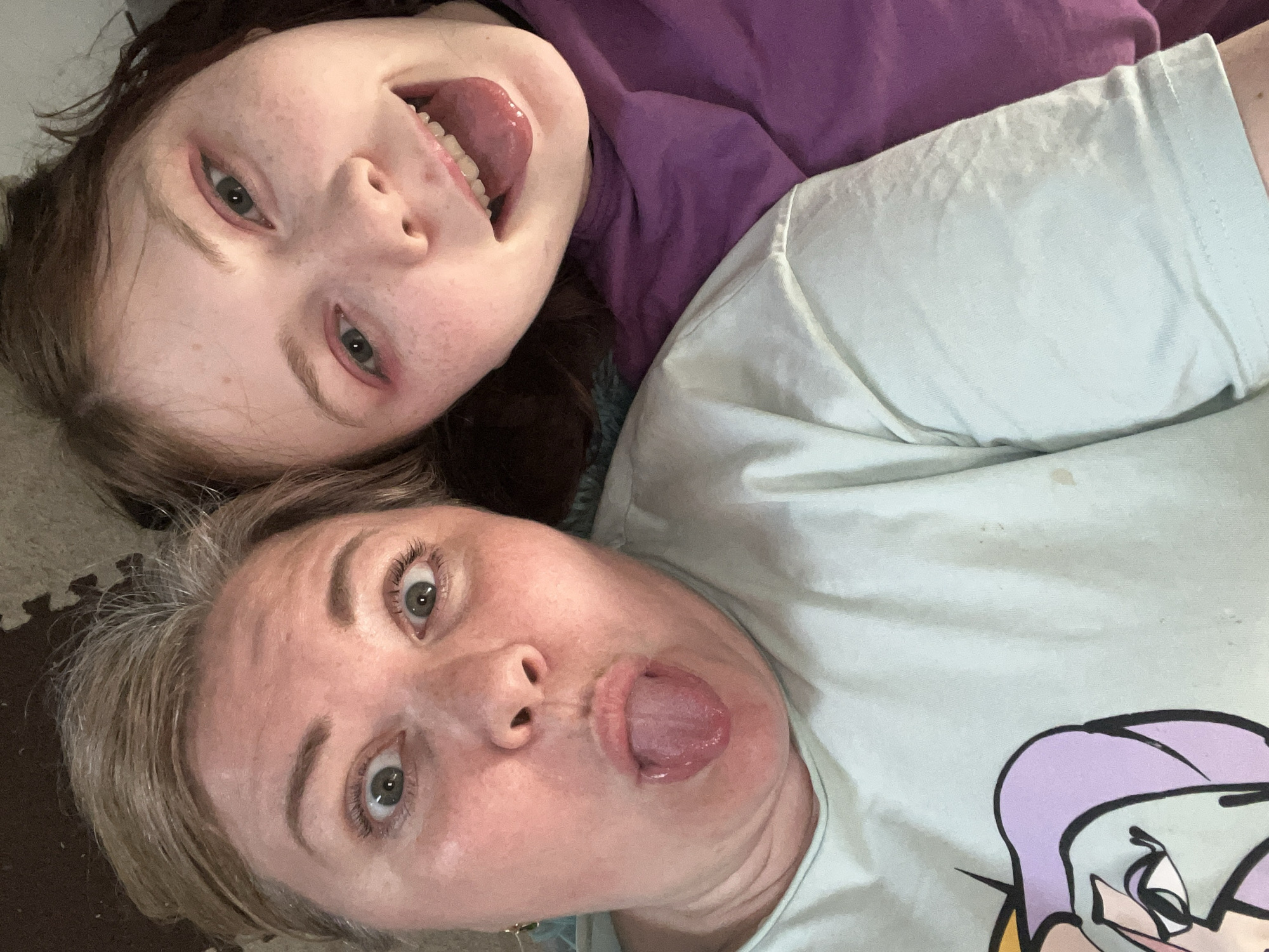 Two individuals, a child and an adult, are captured in a close-up selfie. Both individuals are lying down, facing the camera with their tongues extended. The child, located in the upper part of the frame, has brown hair and is wearing a purple shirt. The adult, positioned below the child, has blonde hair and is wearing a light blue t-shirt with a cartoon graphic. The background shows a mottled beige surface, possibly a floor. There is no indication of the location being Vesyoloye, Russia in the image.