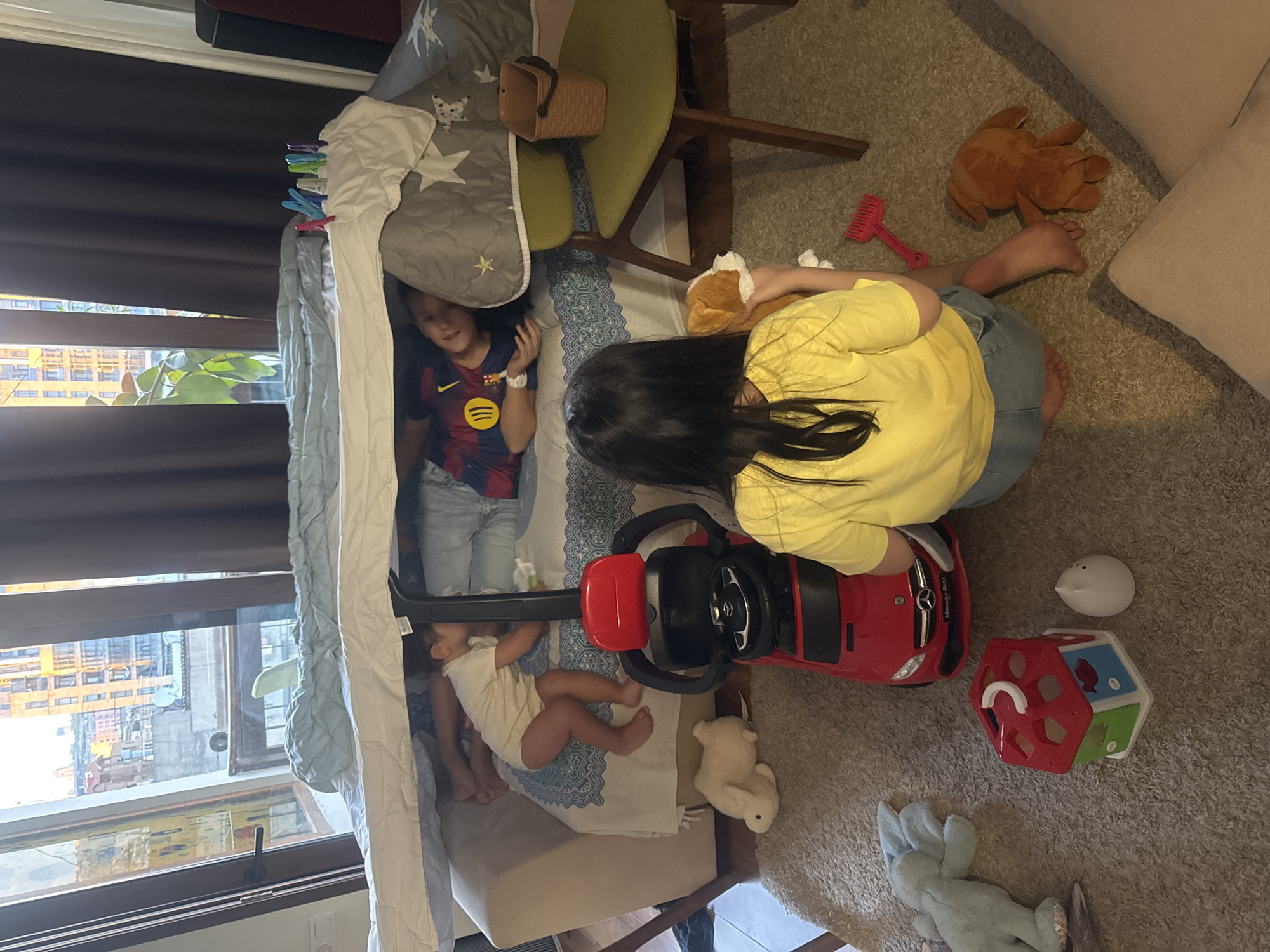 The image depicts an indoor scene, likely a living room, where children are playing. A makeshift fort has been constructed over a sofa, supported by blankets and pillows. Two children are visible within or near the fort. One child, wearing a red and yellow striped shirt, is lying down inside the fort. Another child, wearing a yellow t-shirt and denim shorts, is seated on the floor, facing away from the camera, and holding a stuffed animal. Scattered on the floor are various toys, including a red toy car, a shape sorter house, a stuffed rabbit, and a light blue stuffed animal. A chair with a green seat and wooden legs is positioned to the right of the fort, with a small woven basket on it. The background shows a large window with a view of buildings, suggesting an urban environment. The overall impression is one of children engaged in imaginative play in a home setting. The image does not contain any specific details to determine a location in Yerevan, Armenia.