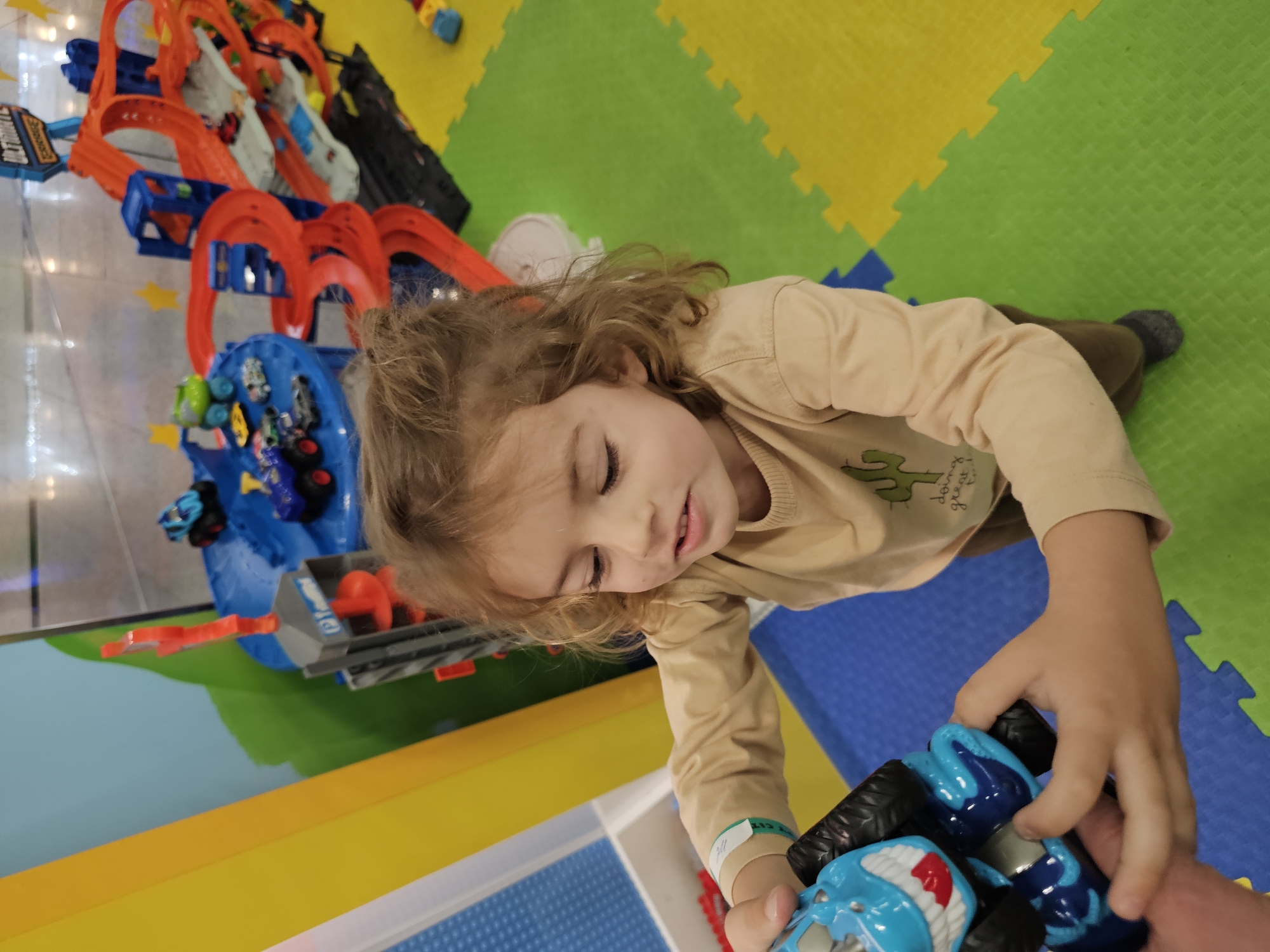 A child with shoulder-length light brown hair wearing a beige long-sleeved shirt and brown pants lies on a colorful play mat. The mat consists of interlocking foam tiles in green, yellow, and blue. The child's eyes are closed, and a slight smile is visible. They are holding a blue and black toy car with both hands. To the left is a toy car track with orange loops and a blue launchpad. Various toy cars are on the track. The scene appears to be in a children's play area located in Khimki, Russia.