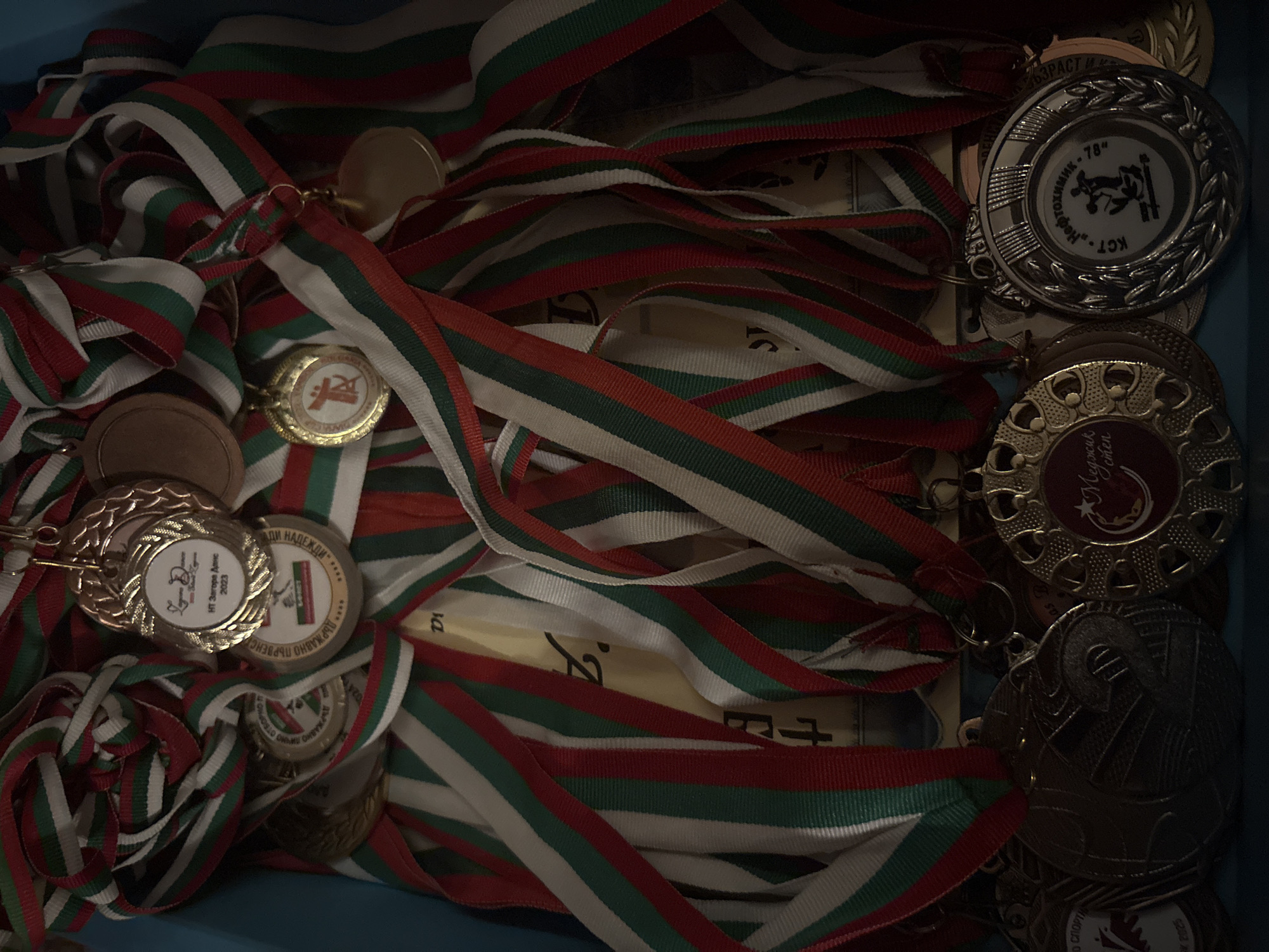 The image is a close-up, high-angle shot of a pile of medals with colorful ribbons. The medals are of various sizes and materials, including gold, silver, and bronze. Many of the medals have text and imagery on them, though they are not all legible. The ribbons are striped in red, white, and green. The medals are clustered together, obscuring much of the background, which appears to be a light blue surface. The lighting is dim, casting shadows on some of the medals.