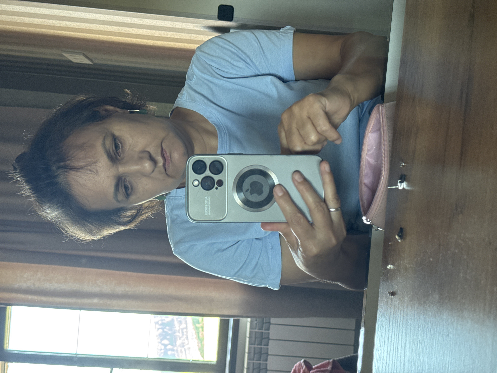 The image is a selfie taken by a woman indoors, reflected in a mirror. The woman has dark brown hair pulled back and is wearing a light blue t-shirt and earrings. She is holding a silver smartphone with a distinctive camera module in her right hand, and her left hand is also visible. In the foreground, a wooden table surface is visible, with small debris and a partially opened pink cosmetic bag. Behind her, a window with a radiator and brown curtains is partially visible on the left, and a wall with vertical stripes and a light switch is on the right. The scene appears to be within a residential or hotel room. There is no discernible information in the image to indicate a specific location in Soloneshnoye, Russia.