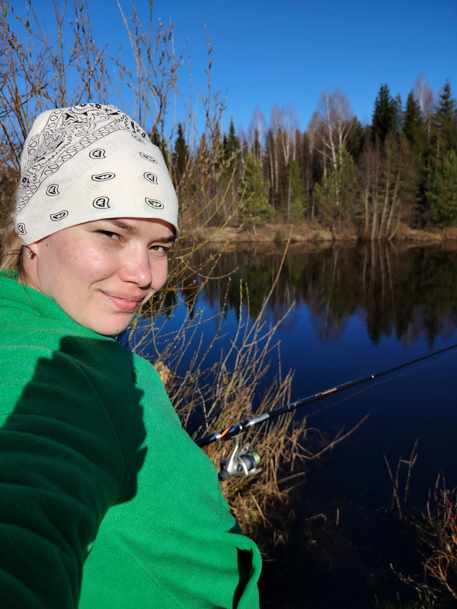 The image shows a woman in Chaikovsky, Russia fishing by a lake or river. She wears a white bandana and a green sweatshirt. Behind her, the water reflects the trees on the opposite bank, which include both bare birches and evergreen firs. A fishing rod extends into the water. The time of year appears to be early spring.