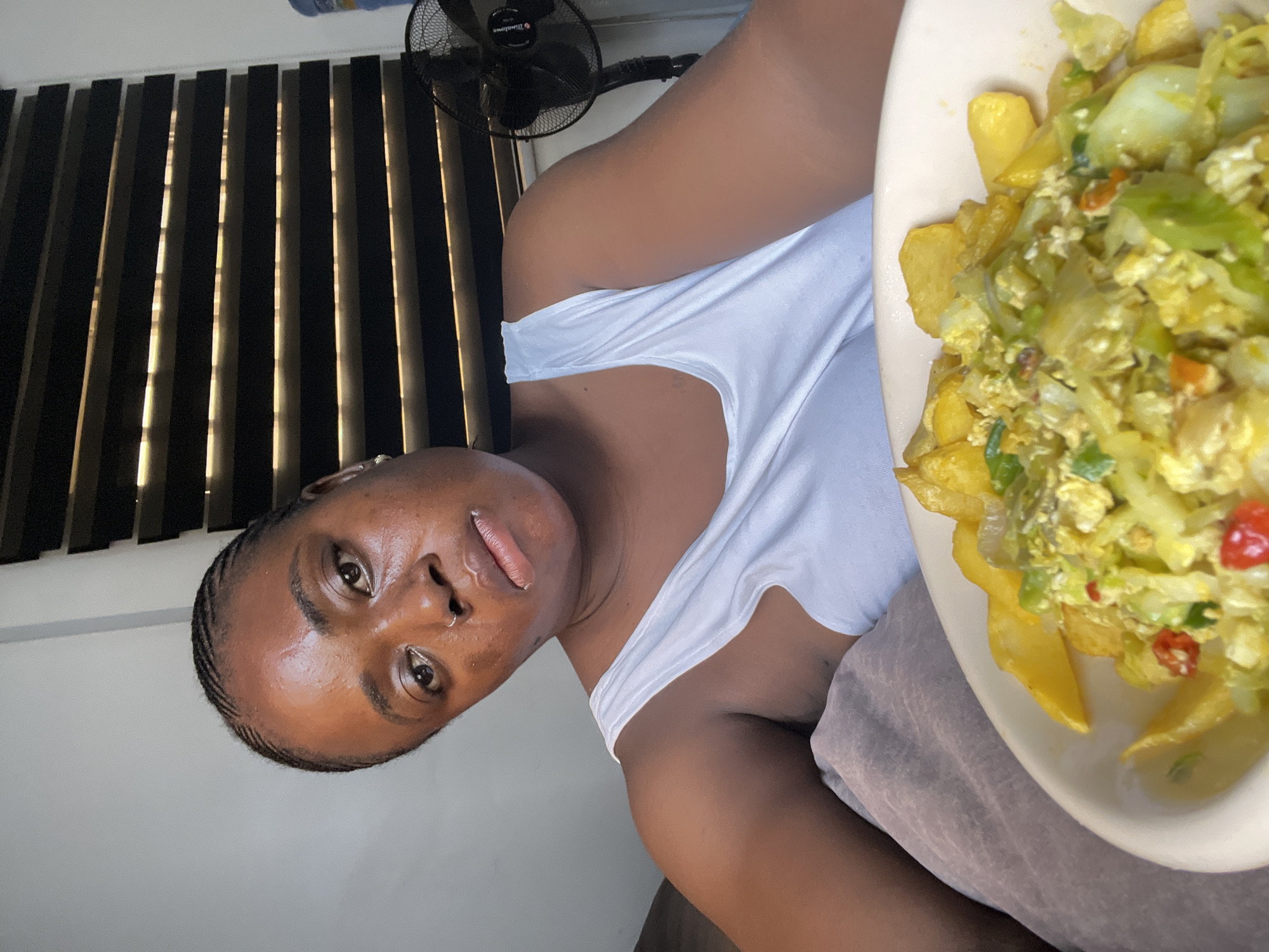 The image shows a young Black woman indoors, with a plate of food to her right. She is wearing a white tank top. Her hair is braided in thin cornrows. She has a nose ring. The background shows vertical blinds and a black fan. The woman's face is turned towards the camera, and she is looking directly at it with a neutral expression. The plate of food contains what appears to be scrambled eggs mixed with vegetables and french fries. The lighting suggests it is daytime. The location context is Abuja, Nigeria.