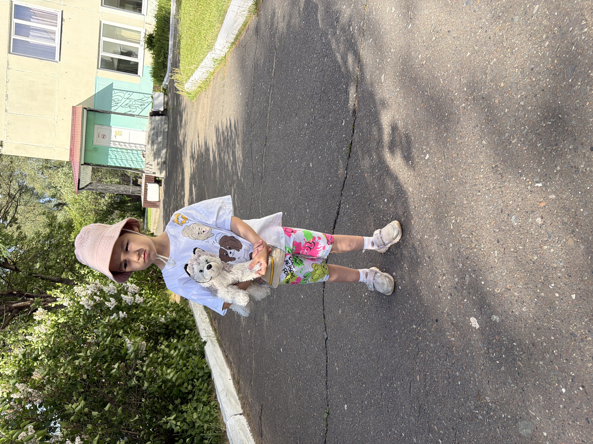 A young child, around 5-7 years old, is standing on an asphalt road next to a sidewalk in Chaikovsky, Russia. The child is wearing a pink bucket hat, a white t-shirt with cartoon dog designs, and shorts with a colorful, floral pattern. They also have on white socks and sandals. The child is holding a teddy bear. To the left of the child is a bush with white flowers. In the background, there is a multi-story building. The day appears to be sunny, as there are shadows cast on the road.