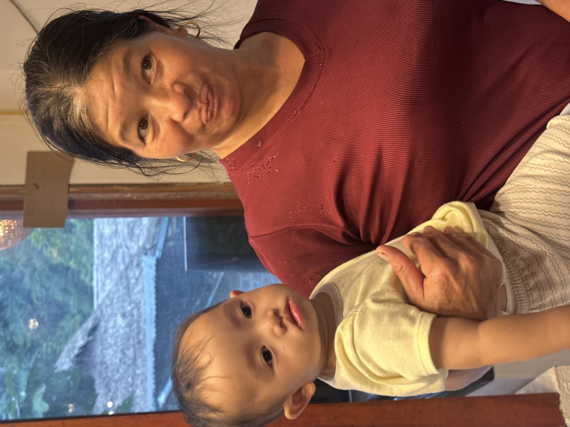 An adult woman holds a baby in front of a window with a thatched roof visible outside. The woman, with dark hair and gold hoop earrings, is wearing a maroon v-neck shirt. She is looking to her right with a slight pout. The baby, wearing a yellow shirt and light-colored patterned pants, is looking to its left with a neutral expression. The setting appears to be indoors, with a hint of natural light coming from the window. The outside view suggests a rural or village environment in Cổ Lũng Commune, Vietnam.