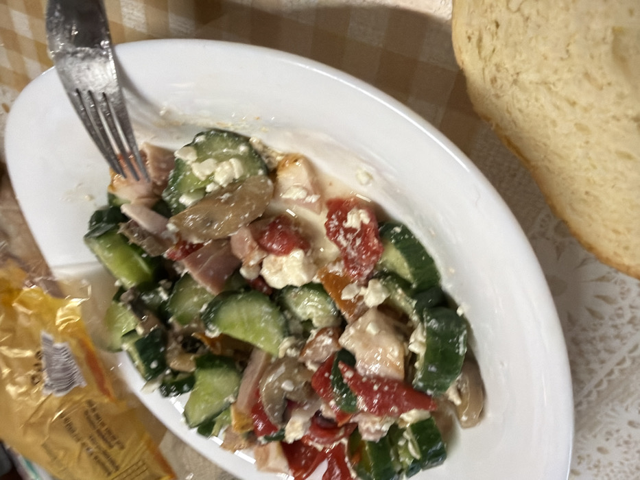 An overhead, slightly angled photograph captures a white oval plate containing a mixed salad, positioned on a patterned tablecloth. The salad consists of thick slices of cucumber, irregular pieces of red bell pepper, crumbled white cheese, light pink processed meat, and sliced mushrooms, all coated in a liquid dressing. A silver dinner fork is partially inserted into the salad on the right side of the plate. A slice of light-colored bread is visible in the bottom right corner of the frame. In the blurry upper left background, a yellow plastic bag with indistinct text is present. The tablecloth features a light-colored checkered pattern in the upper right and a white lace or embroidered pattern in the lower left. No individuals are visible. The specific location within Tarnava, Bulgaria, cannot be determined from the visual evidence provided; the setting appears to be an indoor, domestic environment.
