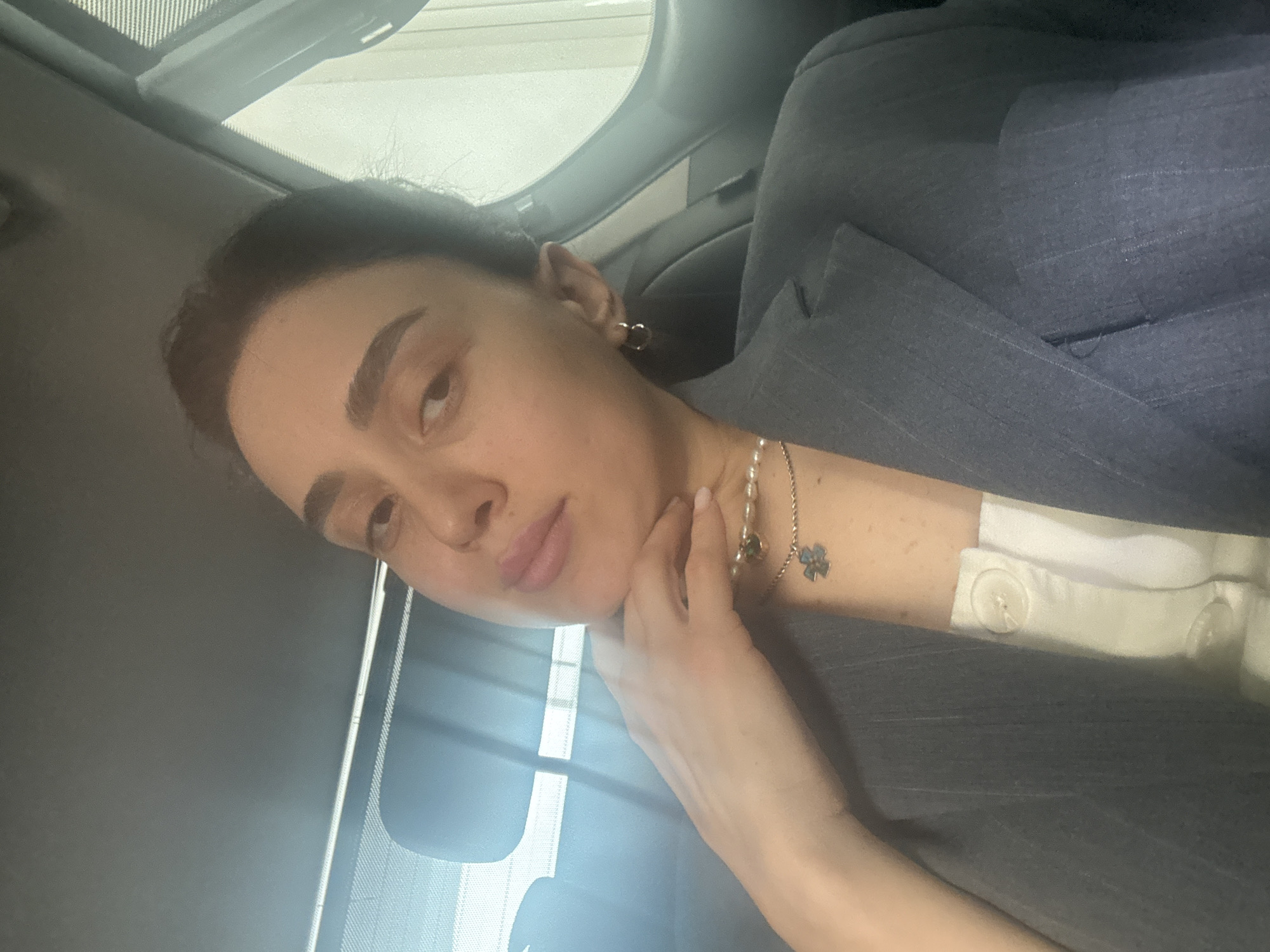 A medium shot photograph shows a light-skinned woman in a vehicle, possibly a car, the scene likely captured during daylight hours. The woman has dark hair pulled back, prominent eyebrows, and is wearing understated makeup. She is adorned with simple gold hoop earrings and a delicate necklace with pearls and a small, possibly clover-shaped pendant. She is wearing a tailored gray pinstripe blazer over a white shirt with buttoned cuffs. Her left hand is raised, with fingertips gently touching her neck. The background features the interior of the vehicle, including a sun visor and a window. The location in Tbilisi, Georgia is not confirmed by the image.