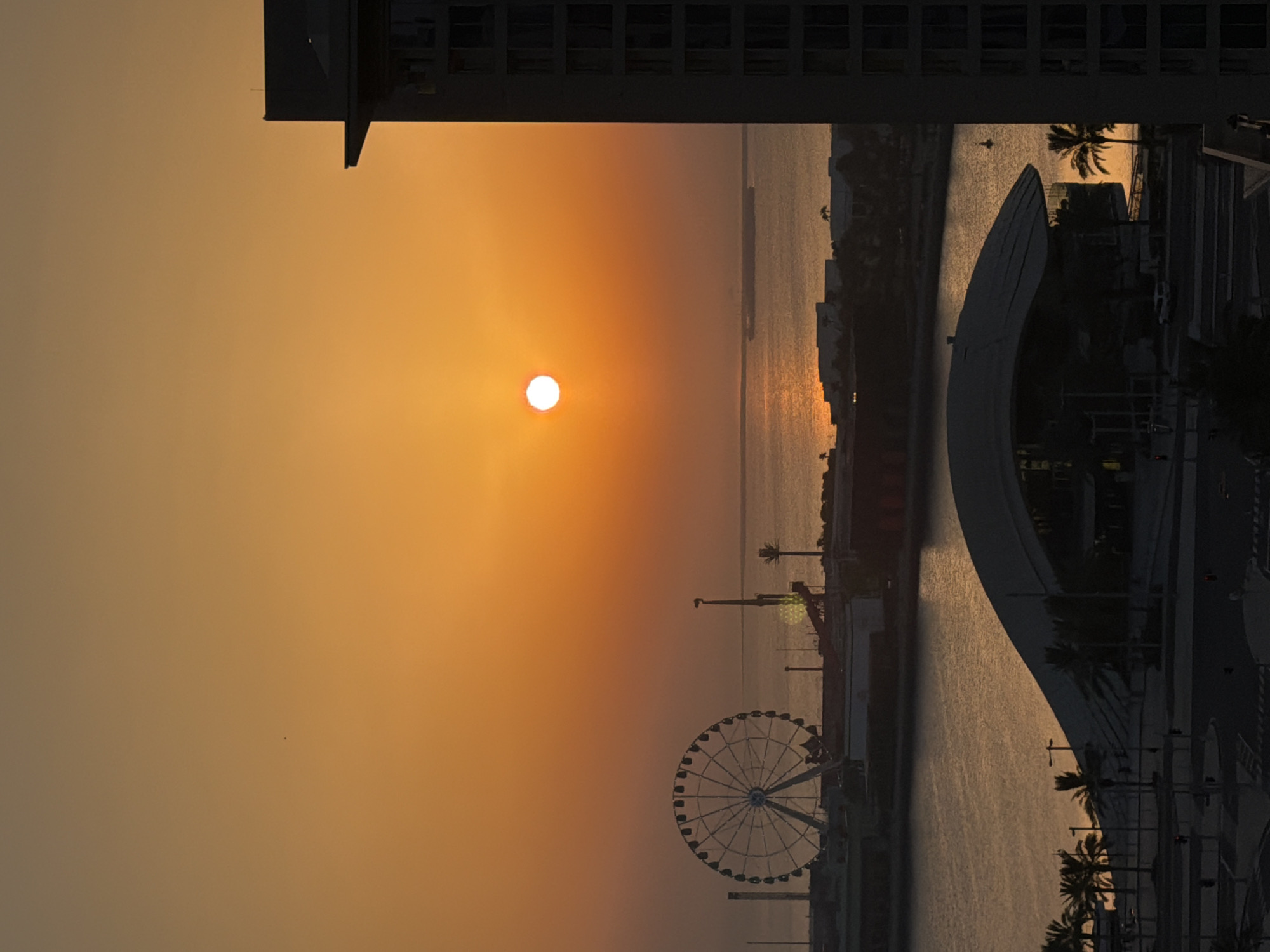 An aerial vertical shot captures a wide view of a city waterfront at sunset. The sky is a vibrant gradient of orange and yellow, with the sun a bright white orb in the upper center. The water reflects the warm hues of the sky. In the distance, a large Ferris wheel stands out against the horizon. Several buildings line the waterfront, including a modern building silhouetted on the right side of the frame. In the foreground, a dark, curved structure, possibly a building or pavilion, dominates the lower part of the image. Palm trees are visible along the waterfront and in the foreground. A ship is seen in the distance on the water. The scene appears to be a developed urban area with recreational facilities.