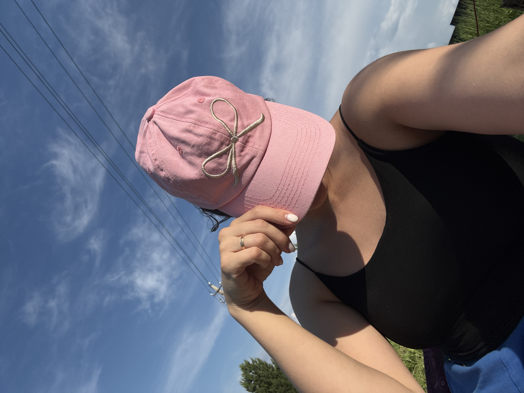 The image is a close-up, low-angle, outdoor selfie taken on a sunny day with a bright blue sky and wispy white clouds. In the foreground, a person is wearing a pink baseball cap with a gold embroidered bow on the front. The person is also wearing a black tank top. Their left arm is bent at the elbow, and their hand is raised, touching the brim of the cap, partially obscuring their face. A silver ring is visible on their ring finger. In the background, several dark power lines stretch horizontally across the sky, with a utility pole visible on the right side. To the left, a portion of a green tree is visible. The lower right corner shows a hint of green grass and a dark, horizontal structure, possibly a fence. The overall setting appears to be an open, possibly rural or suburban, area with overhead power lines. There is no specific indication of the city of Doksha, Russia, within the visual information provided.