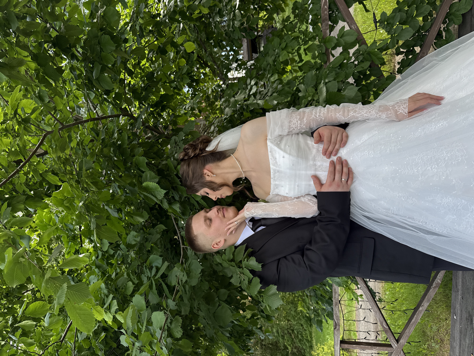 A medium shot captures a newlywed couple embracing outdoors, positioned in front of lush green foliage. The groom, wearing a black suit with a white shirt and black tie, gently touches the bride's cheek. The bride, adorned in a white off-the-shoulder wedding gown with lace sleeves and pearl embellishments, leans into his touch. Her hair is styled in an updo, and she wears a delicate pearl necklace. They are standing on a wooden deck with a decorative railing and string lights. The background is dominated by the dense leaves of a large tree, with hints of grass and a wooden structure visible through the branches. The scene suggests a serene and romantic moment captured in an outdoor wedding setting, possibly in a park or garden area in Votkinsk, Russia, given the presence of lush vegetation and traditional wooden architectural elements.