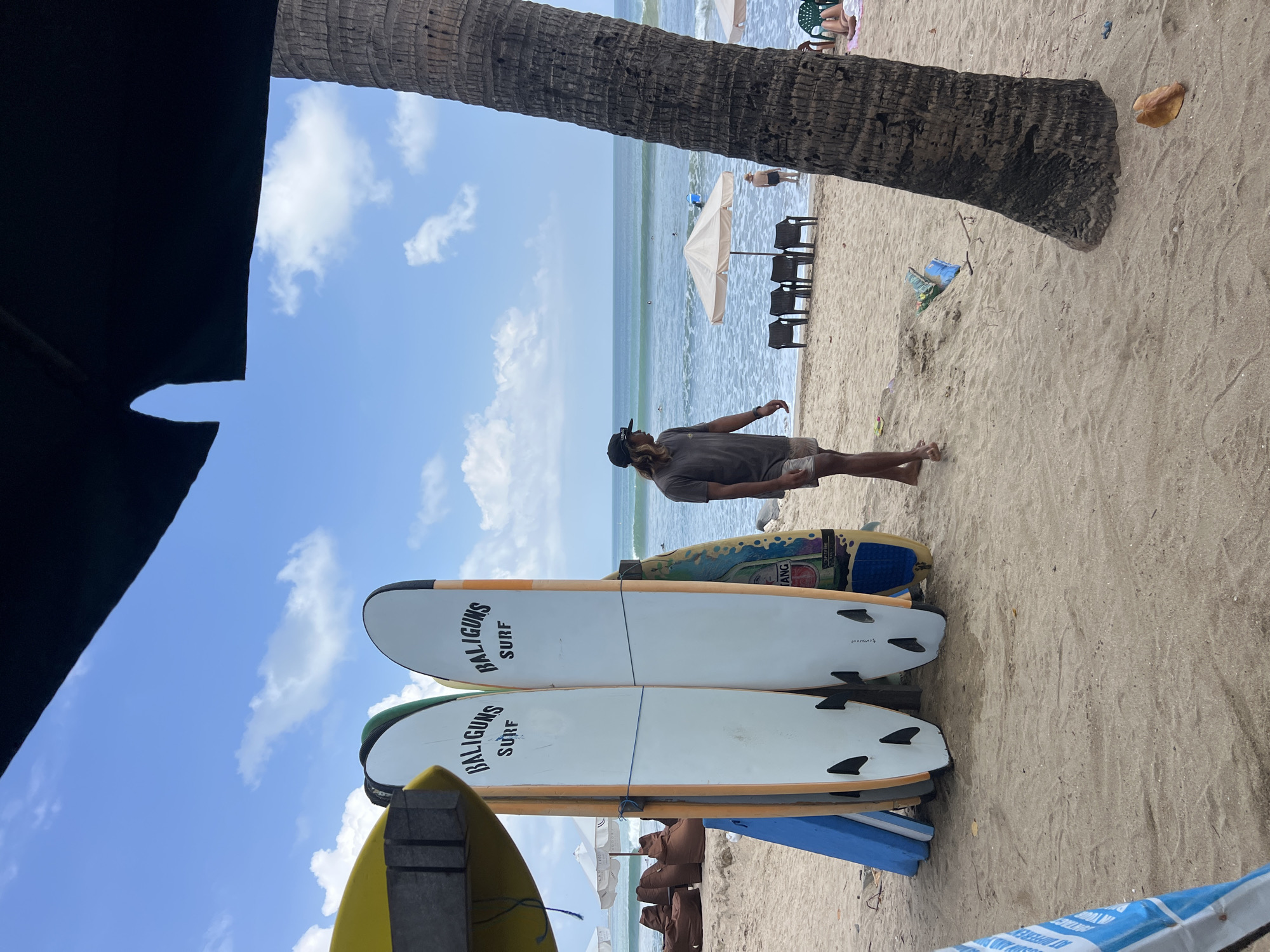 A man with long hair, wearing a dark t-shirt and shorts, stands on a sandy beach in Kuta, Indonesia. He is looking towards the ocean, which has moderate waves and several surfers. Behind him, a rack of surfboards is propped up on the sand, some with the text "BALIGUNS SURF" visible. A tall palm tree trunk is on the right side of the image. Beach chairs and umbrellas are set up further down the beach, with other beachgoers visible. The sky is blue with scattered clouds.
