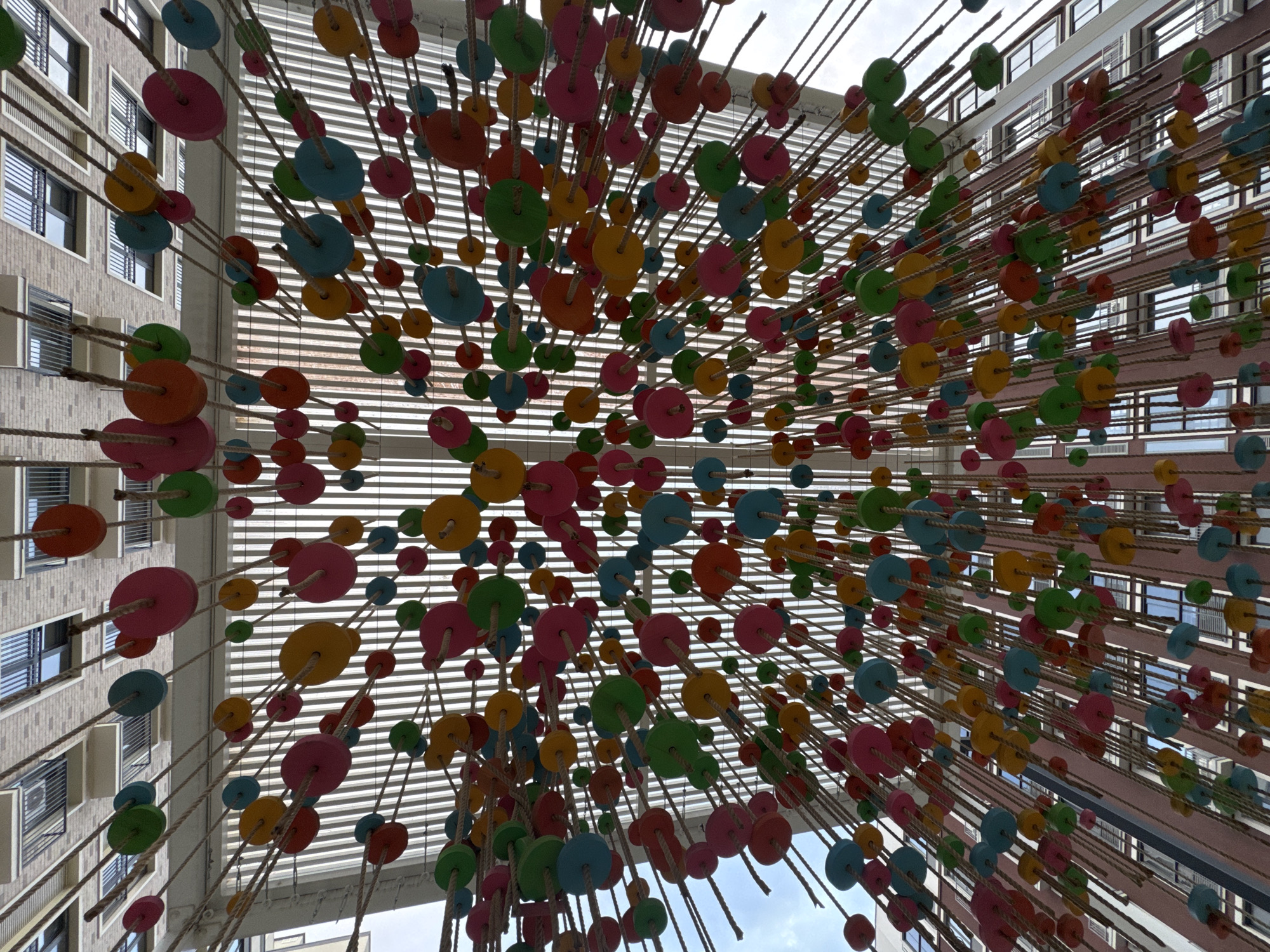 The image is an upward-angle shot of a colorful mobile installation hanging from a building's facade in Izhevsk, Russia. The mobile consists of numerous circular, colored discs in shades of pink, orange, yellow, green, and blue, suspended at varying lengths by ropes. These discs hang against a backdrop of a light-colored, striped roof structure and the windows and balconies of multi-story buildings. The sky is visible in the upper left corner. There are no people or animals visible in the image.