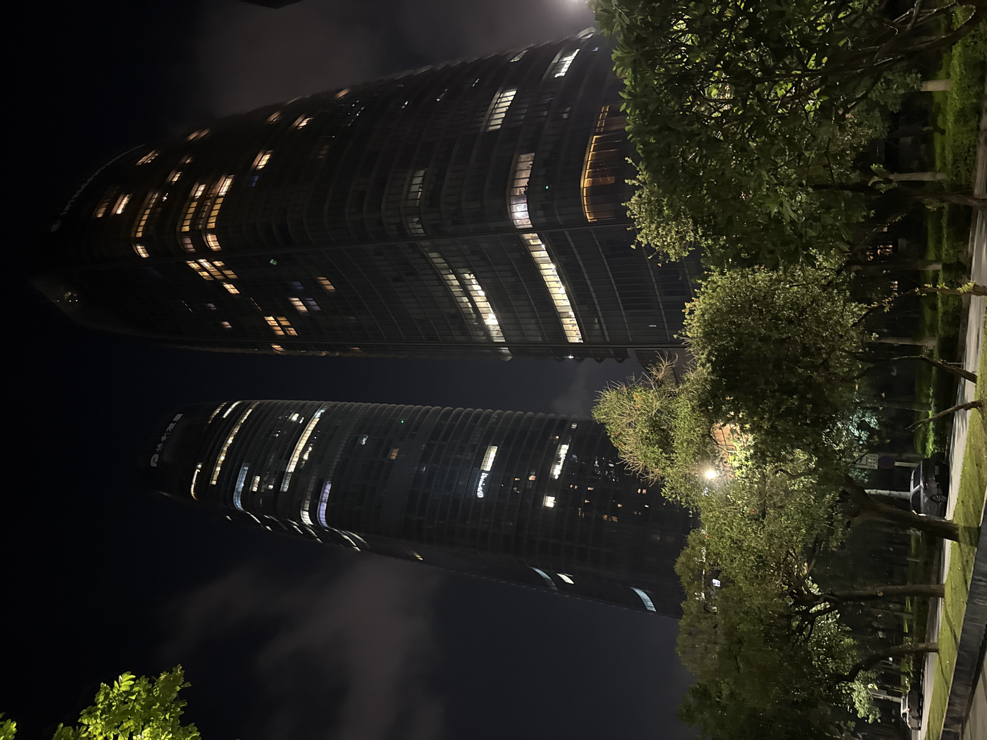 The image shows a night-time scene of two modern skyscrapers in Haizhu District, China. The buildings are tall, with a dark, glass facade illuminated by numerous lit windows. The skyscraper on the left appears to have a slightly curved design, while the one on the right is more rectangular. Trees and foliage line a paved area in the foreground, with some trees obscuring the lower portions of the buildings. A street with parked cars is visible to the left of the trees, and a lamppost is also present within the treeline. The sky is dark with some cloud cover, and the overall lighting suggests an urban environment at night.
