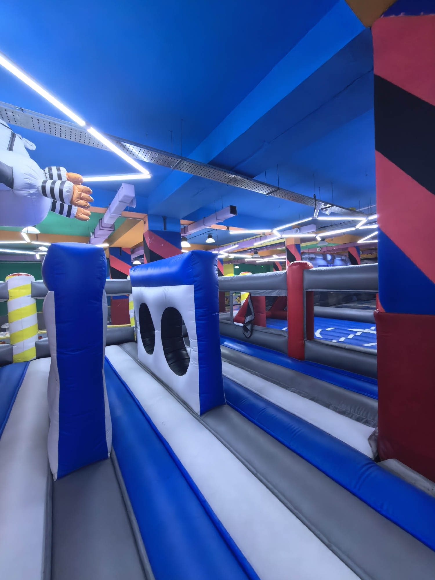 The image captures an indoor inflatable playground, likely located in Fergana City, Uzbekistan. The space is characterized by vibrant blue ceilings with angled fluorescent lighting fixtures, creating a dynamic and illuminated atmosphere. Large inflatable structures, primarily in white, blue, and grey, dominate the foreground and midground, forming various play elements such as walls, tunnels, and possibly obstacle courses.
A prominent feature is a large, white inflatable figure with orange and black striped hands suspended from the ceiling in the upper left of the frame. In the foreground, there are large blue and white inflatable structures with cut-out openings, resembling entrances or windows. Adjacent to these, a grey and blue inflatable barrier runs through the midground, suggesting a pathway or a separated play zone. In the background, more inflatable structures and what appear to be padded red and black columns are visible, contributing to the overall colorful and playful environment. The floor beneath the inflatable structures is not fully visible but appears to be padded or a soft surface. No individuals are present in the scene.