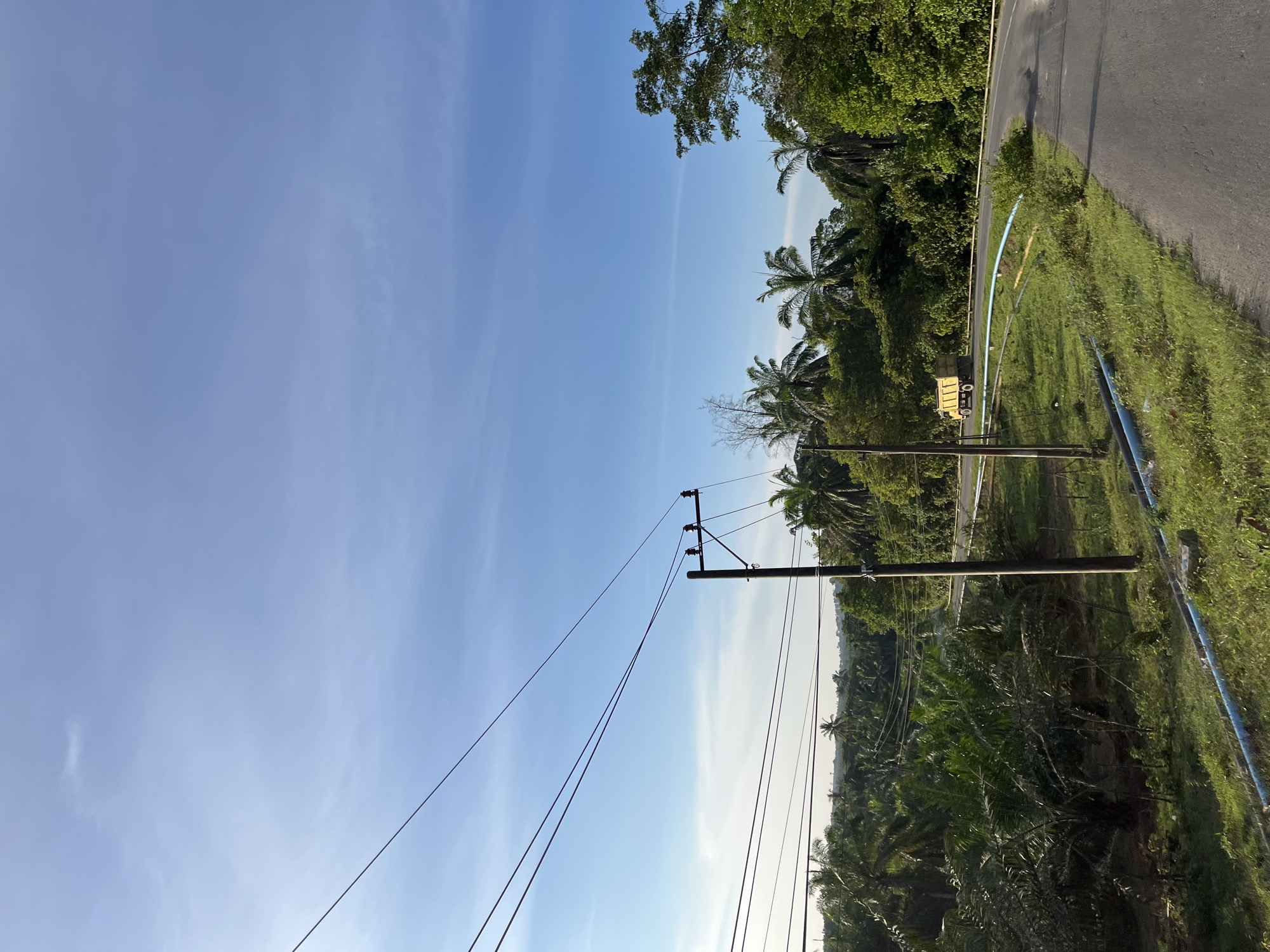This image depicts a scenic roadside view in Sukaramai Dua, Indonesia, under a clear blue sky with wispy clouds. The foreground is dominated by a power pole with multiple wires stretching across the frame, connecting to insulators. To the right of the pole, a paved road curves gently through lush green vegetation, primarily consisting of palm trees and other dense foliage. A bright yellow dump truck is visible on the road, navigating the curve. Blue pipes run along the edge of the grassy verge next to the road. The background shows a rolling landscape covered in trees, suggesting a rural or semi-rural setting. The light indicates it is likely morning or late afternoon, with the sun casting a warm glow. There are no people visible in the scene.