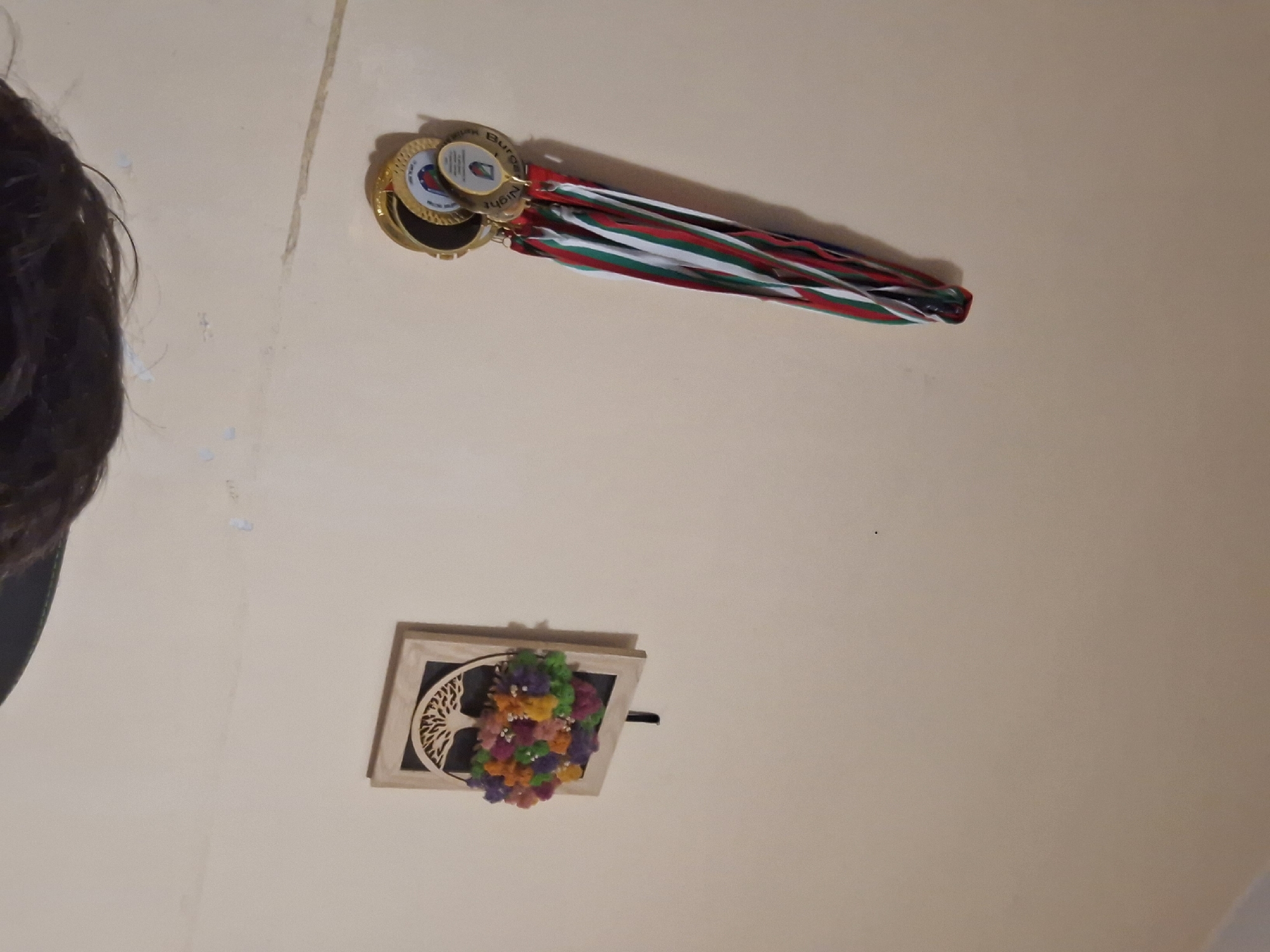 The image shows a view of a ceiling. Attached to the ceiling are medals with ribbons in the colors of the Bulgarian flag (white, green, and red), and a framed decorative item with a tree-of-life design, adorned with colorful artificial flowers. It is not possible to determine the exact location within the city of Burgas, Bulgaria, from the image alone.