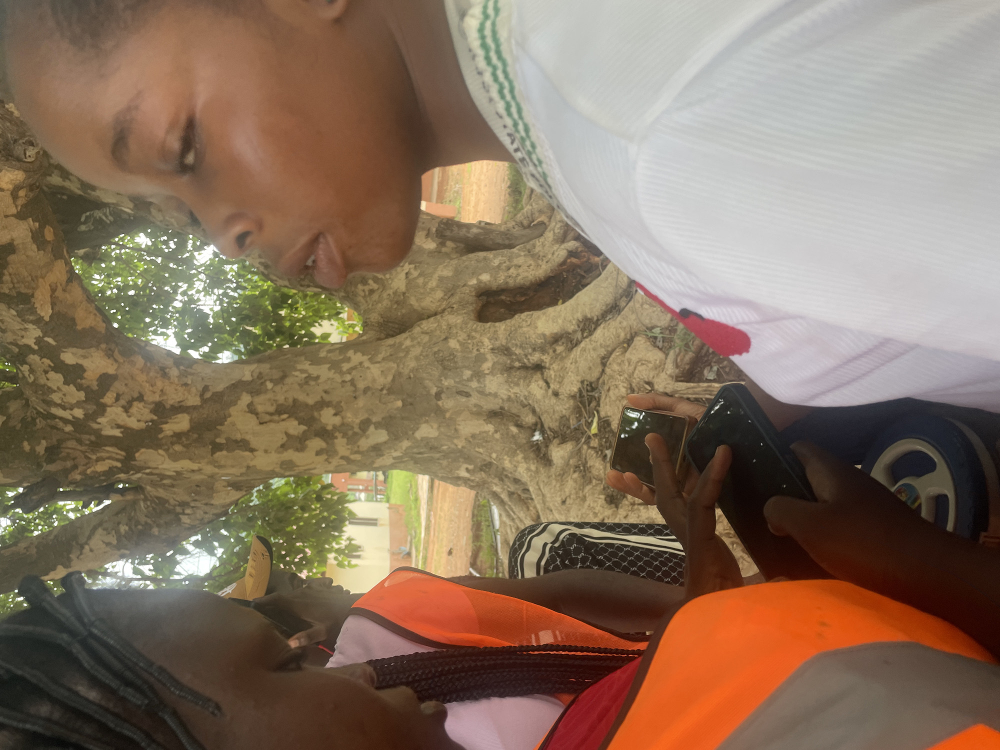 In Lokoja, Nigeria, a group of individuals are gathered outdoors near a large tree with exposed roots. Two people are prominently featured in the foreground. One person, on the left, is wearing an orange safety vest over a pink garment, and their hair is in braids. They are holding and looking at a smartphone. Another person, on the right, is seen in profile, wearing a white shirt with green and white trim around the collar. Their facial features suggest they are speaking. The background reveals the textured bark of a mature tree, its extensive root system visible, and beyond that, glimpses of buildings and foliage under a bright sky. The overall scene appears to be an informal gathering or meeting outdoors, possibly in a park or a public space.