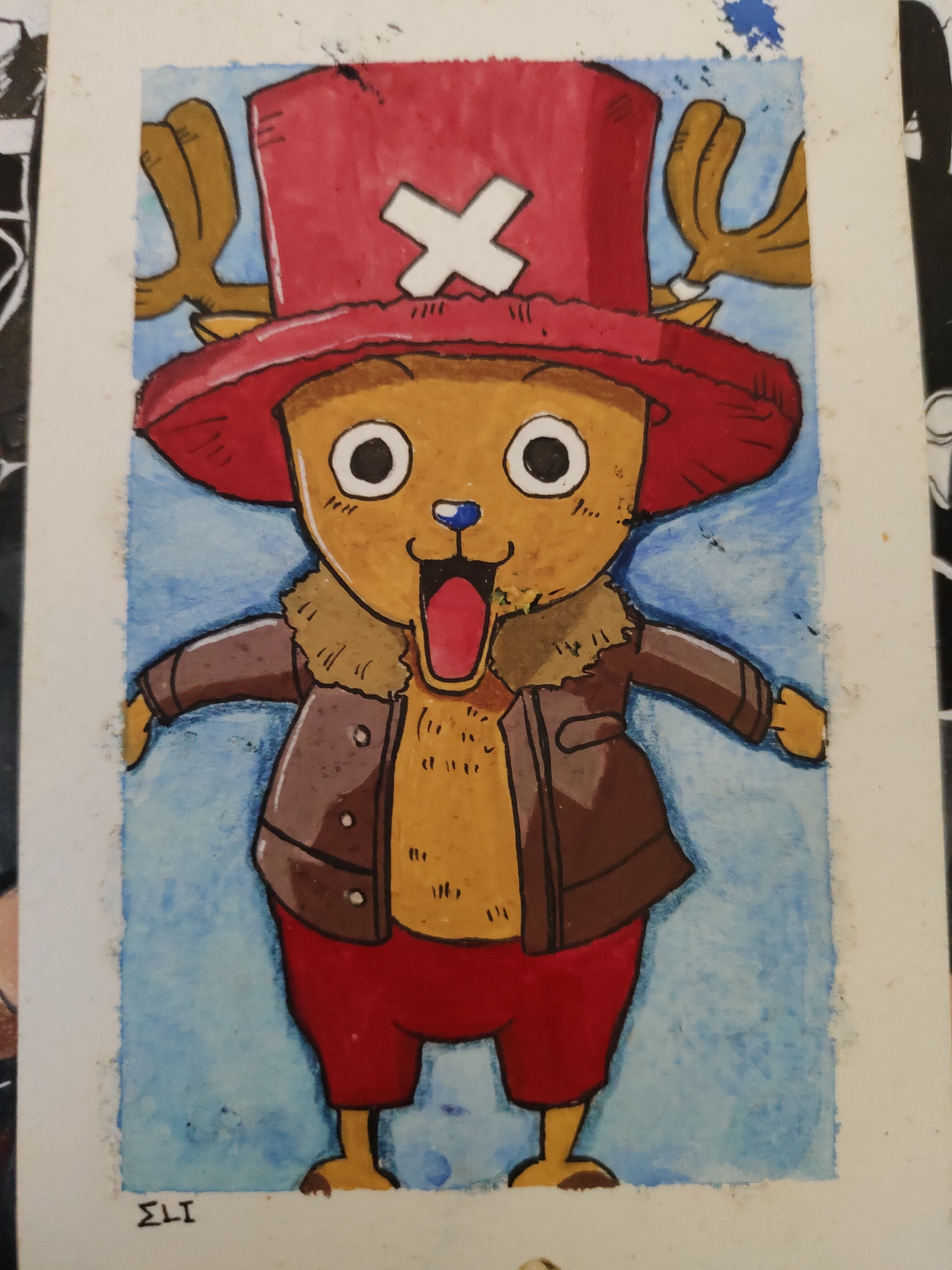 The image displays a watercolor drawing of the character Tony Tony Chopper from the *One Piece* series. The character is depicted with light brown fur, large white eyes with black pupils, a blue nose, and an open mouth revealing a red interior and tongue. Chopper wears a prominent red top hat marked with a white 'X' symbol, with two brown antlers extending from behind the hat. The character is dressed in a brown, long-sleeved jacket with a light brown fur collar, and red shorts or pants. The background of the drawing is a uniform light blue. The artwork is presented on white paper, with the edges of the paper visible, exhibiting some textural imperfections and minor smudges. A black signature, "ΣLI", is present in the bottom-left corner of the drawing. The image itself is a photograph of this drawing. The image provides no discernable information regarding its location in Davao City, Philippines, or any other specific geographical area.