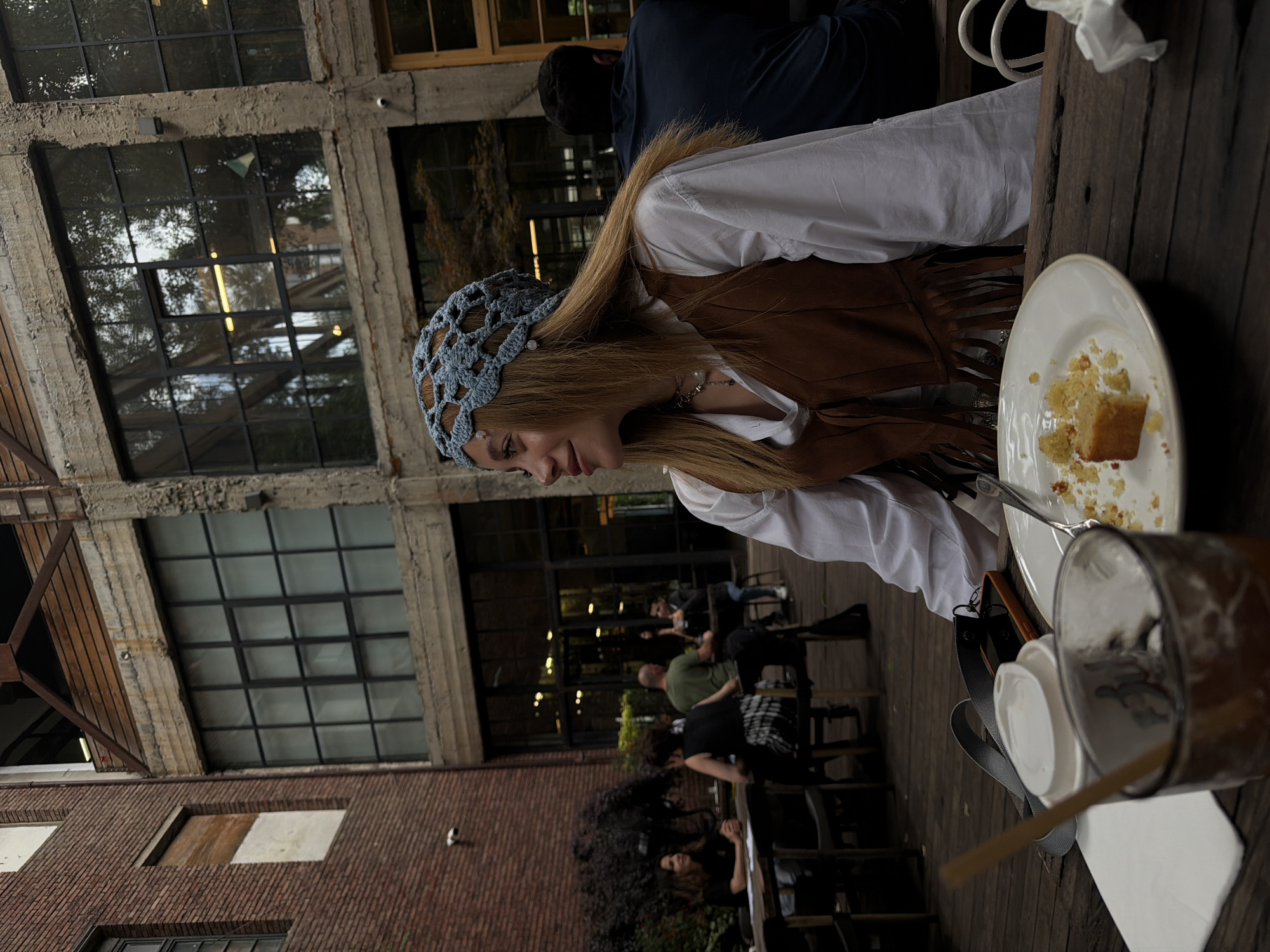 The image captures an outdoor seating area of a cafe, likely located in the Vera district of Tbilisi, Georgia. The architectural style suggests a repurposed industrial building with exposed brick and large windows. A young woman is seated at a table in the foreground, wearing a white shirt, a brown fringed vest, and a blue crochet head covering. She is looking to her left, with a partially eaten piece of cake and a drink on the table in front of her. Other patrons are visible in the background, seated at tables, with some engaged in conversation. The overall atmosphere appears relaxed and casual, typical of an urban cafe setting.