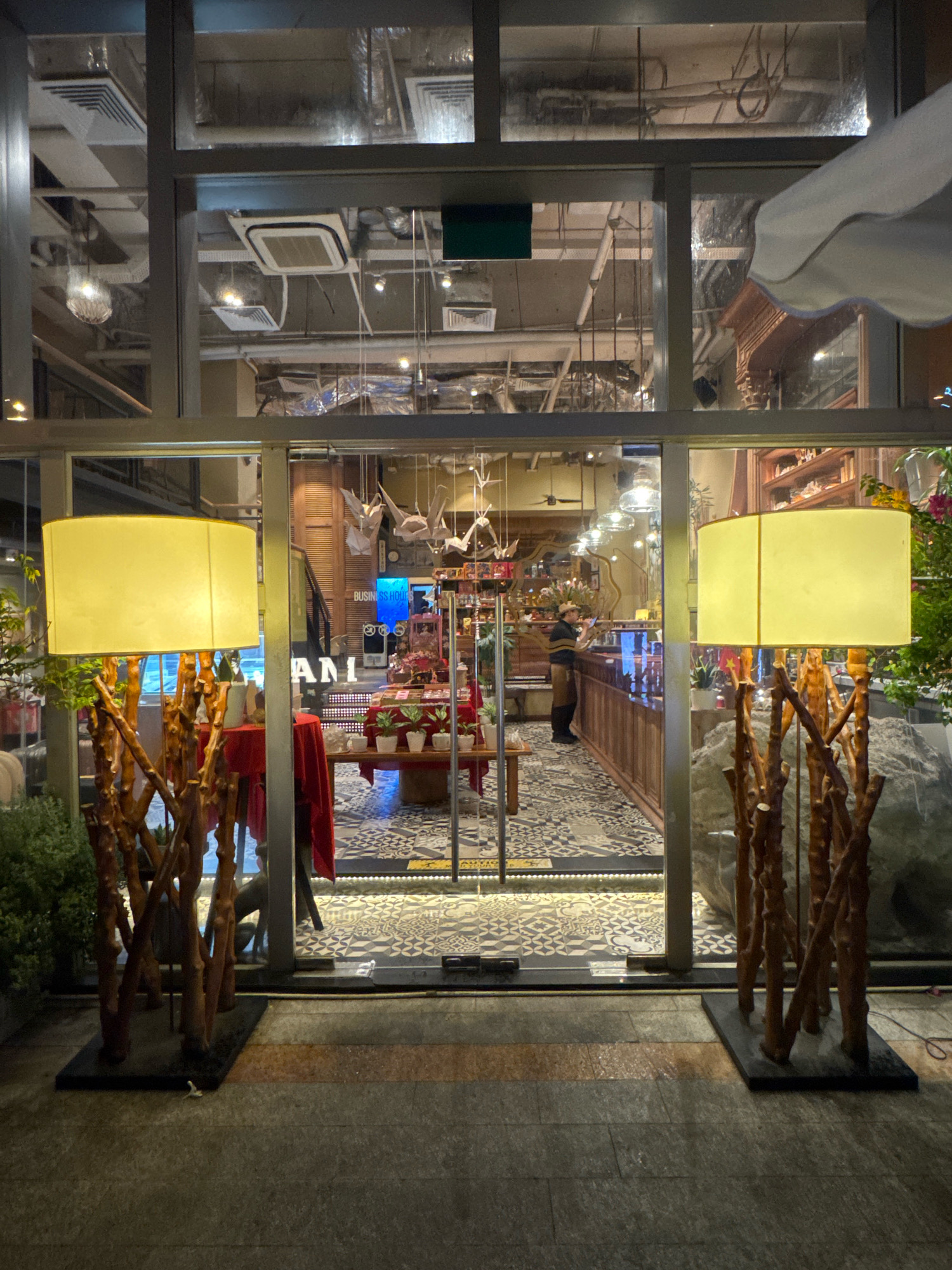 The image shows the exterior of a storefront in Nha Trang, Vietnam, at night. Two large lamps with yellow shades stand on either side of a glass entrance. The lamps have bases made of intertwined branches. Through the glass, the interior of the store is visible, featuring displays of merchandise, shelves, and a counter. A person wearing a brown apron and a hat stands behind the counter, facing away from the camera. Decorative elements like paper cranes are suspended from the ceiling inside. The floor outside the store is paved with dark, rectangular stones, and the floor inside has a patterned tile design. The scene suggests a retail environment, possibly a boutique or gift shop.