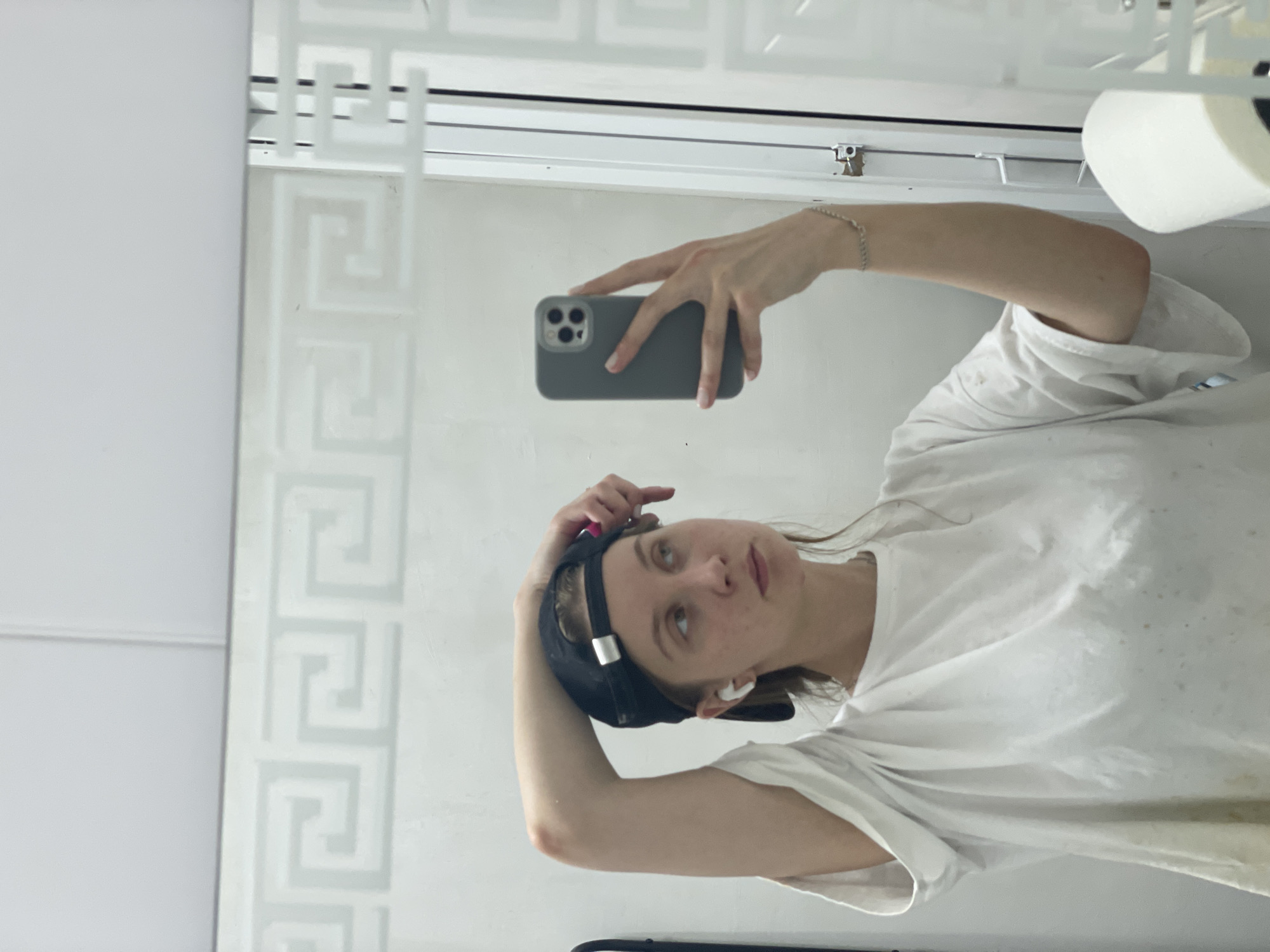 A young woman with light brown hair, wearing a black baseball cap, a white t-shirt, and white wireless earbuds, takes a mirror selfie. She is holding a grey smartphone in her right hand, angled to capture her reflection. Her left arm is bent at the elbow, with her hand resting on the back of her head. The background appears to be a bathroom, with a mirror featuring a decorative geometric pattern. A roll of toilet paper is visible on the right side of the frame. The woman's expression is neutral as she looks into the mirror. The image does not contain any specific details that place it in Chaikovsky, Russia.