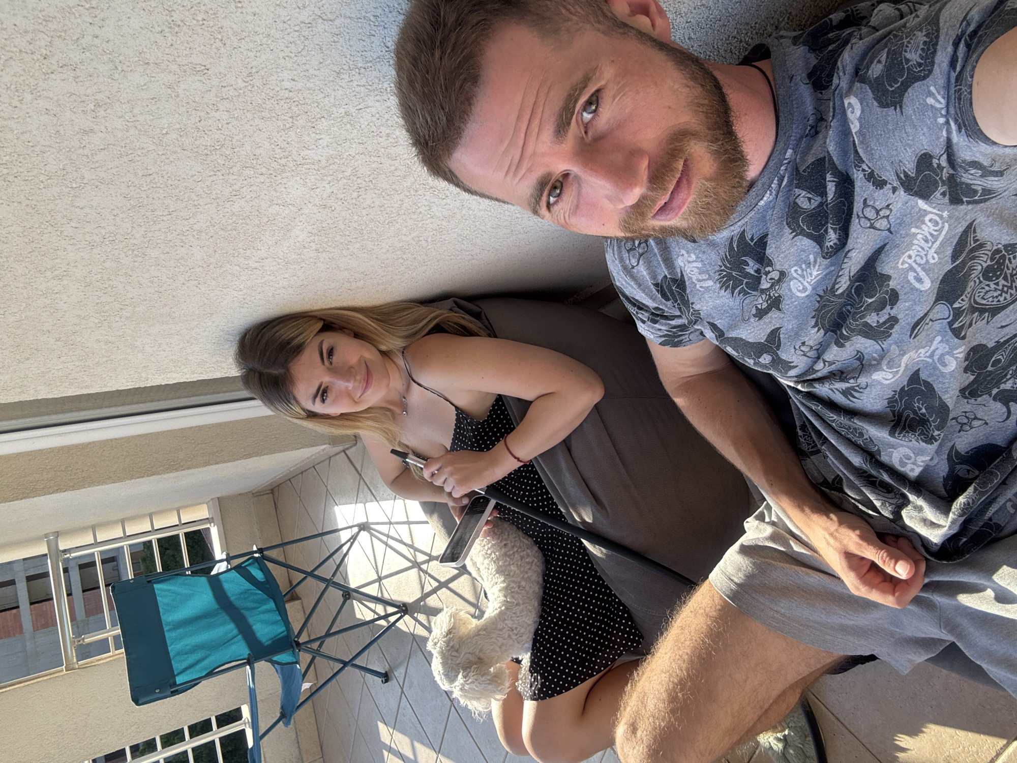 The image captures two individuals and a dog on a balcony in an urban setting, described by the user as Budva, Montenegro, under late afternoon sunlight. A light-colored, textured wall is behind them.
A female individual is seated on a grey bean bag chair against the wall. She has blonde hair and wears a black polka-dot spaghetti strap dress. She holds a white smartphone in her left hand and the black stem of a hookah to her mouth with her right hand. A small, white, curly-haired dog is resting on her lap, facing left. She looks directly at the camera with a slight smile. A delicate silver necklace and a red bracelet on her right wrist are visible.
A male individual is positioned on the tiled floor in the foreground, leaning against the same wall. He has short brown hair and a beard, and wears a grey t-shirt with dark patterned animal graphics and text, and grey shorts. He looks directly at the camera with a subtle expression, his tongue slightly visible between his lips. His right arm rests on his thigh.
To the left, an empty teal folding camping chair stands on the tiled balcony. The balcony features a metal railing. In the background, an unfinished multi-story building constructed of concrete and red brick is visible beyond the railing, alongside other urban structures and some distant greenery. Strong, warm sunlight from the right casts prominent shadows across the beige-tiled floor.