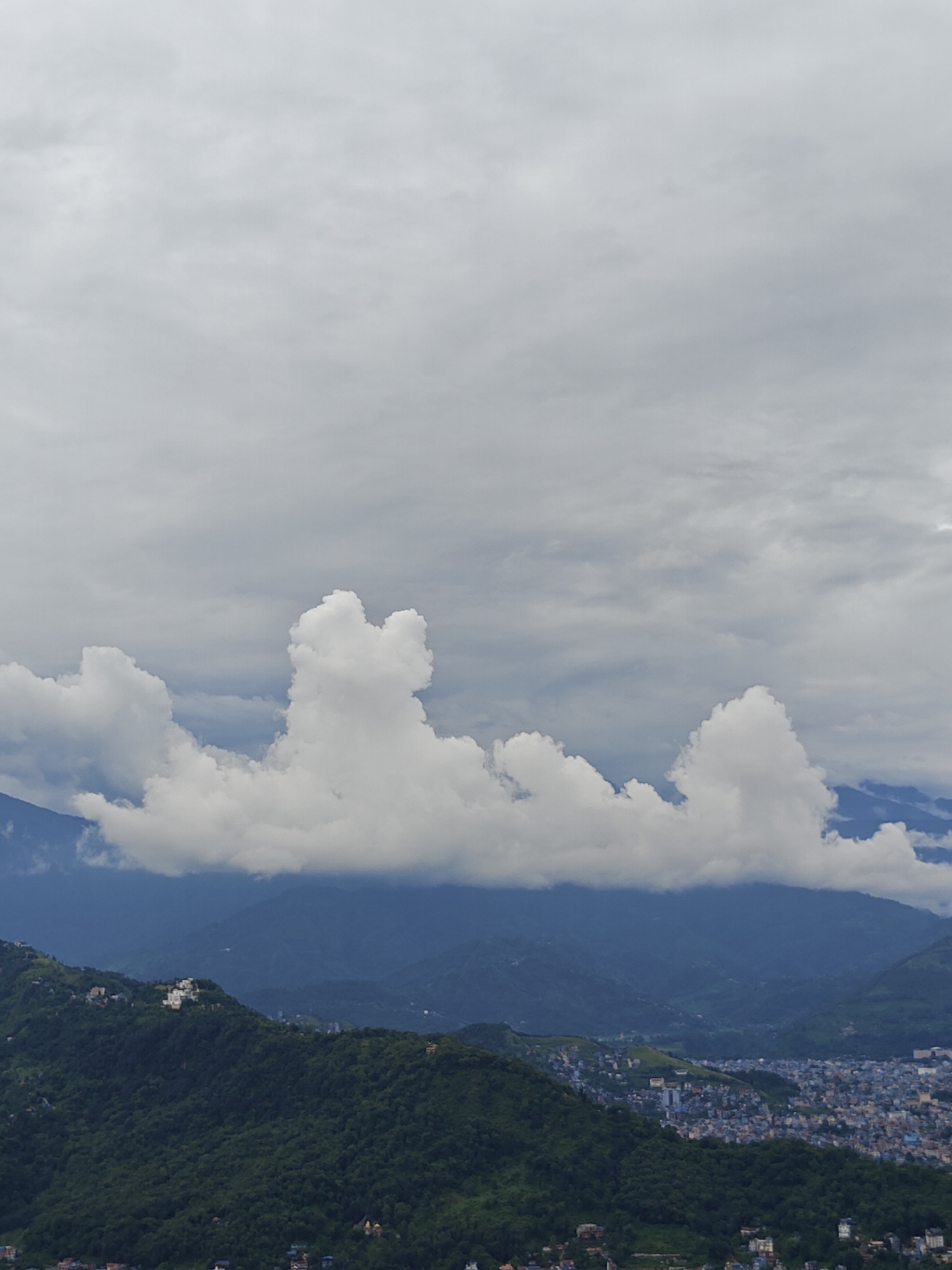 This is a high-angle aerial shot of Pokhara, Nepal, under a cloudy sky. The foreground is dominated by lush green, tree-covered hills dotted with white buildings. A city sprawls across the mid-ground, characterized by a dense collection of low-rise buildings in shades of blue, gray, and white. In the background, a range of hazy blue mountains rises, partially obscured by a band of white clouds that stretch across the horizon. The clouds are illuminated from above, creating a bright, diffused light that casts a soft glow on the landscape. The overall impression is of a serene, overcast day in a mountainous city.