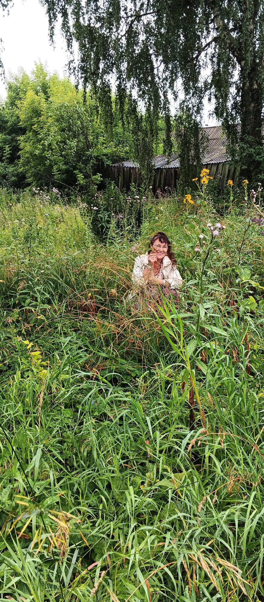 The image depicts a woman kneeling in a dense field of tall grass and wildflowers. She is wearing a light-colored jacket over a patterned dress and has dark hair. The woman is smiling and holding a small flower or plant in her hands, appearing to be examining it. Behind her, a wooden fence runs horizontally, and beyond that, a dilapidated building with a corrugated metal roof is partially visible. The surrounding vegetation is lush and green, with scattered yellow and purple flowers. The scene is set in a rural environment, consistent with a location in the village of Podgornoye, Russia. The lighting suggests it is daytime, possibly overcast, given the soft shadows.