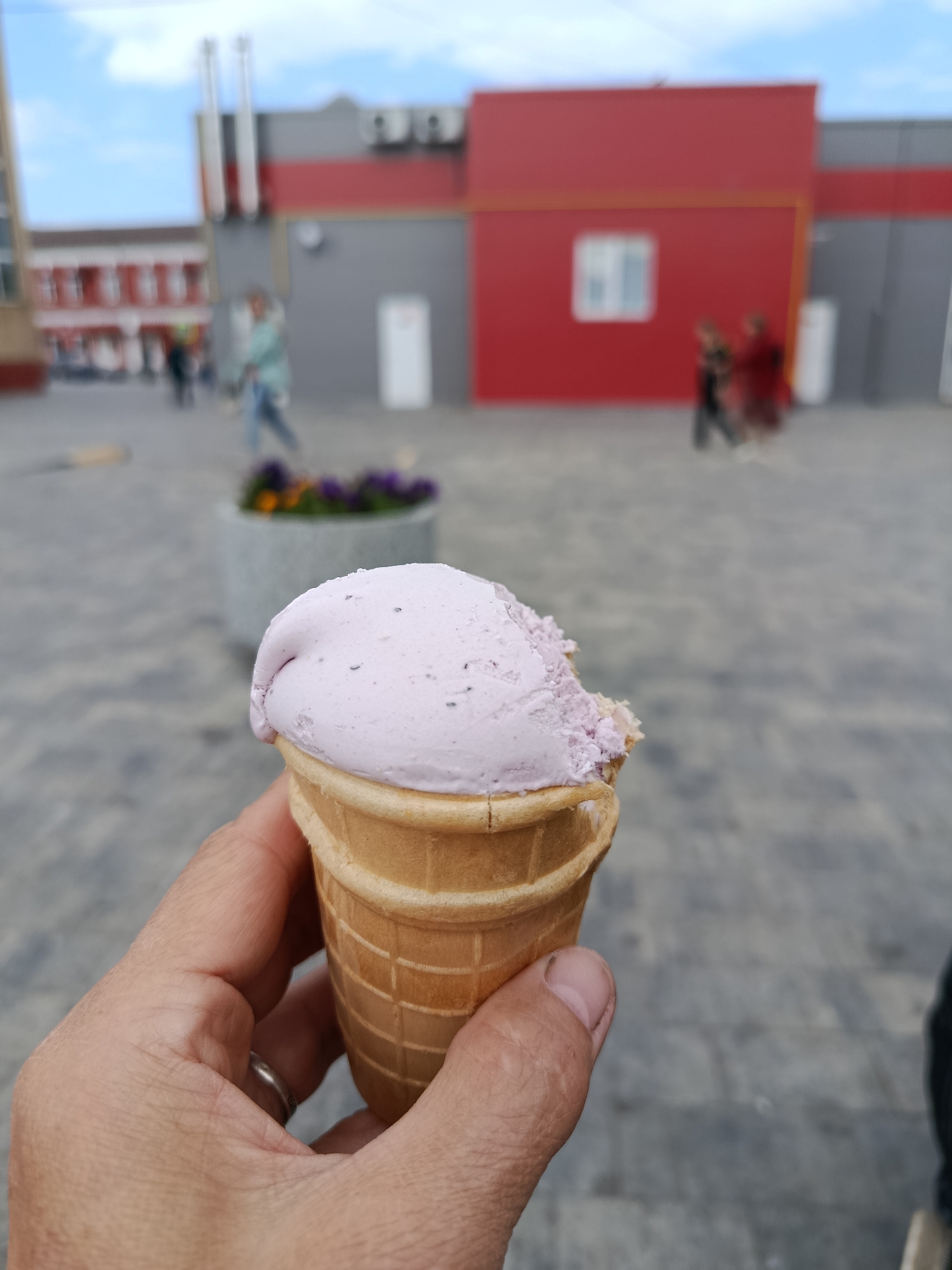 The image captures a close-up of a hand holding a pink ice cream cone with a bite taken out of it. The cone is a light brown waffle cone. The hand is holding the cone from the bottom, with a silver ring on the ring finger. In the background, a blurred urban scene is visible. There is a gray pavement, a concrete planter with purple and yellow flowers, and several buildings with red and gray facades. The sky above is partially cloudy. The people in the background are indistinct due to the blur. This scene appears to be in an outdoor public space, possibly a square or a pedestrian area, within the city of Orekhovo-Zuyevo, Russia.