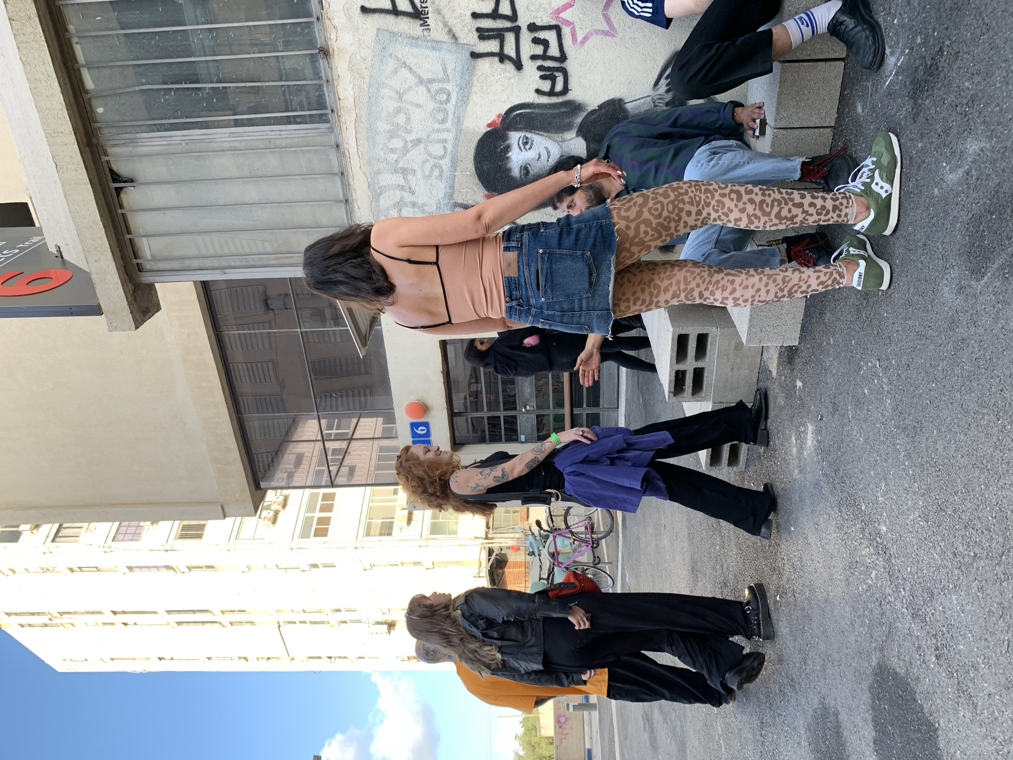 In Tel Aviv, Israel, a group of people are gathered in an outdoor urban setting. Three individuals are visible: one is seated on a concrete structure with graffiti on the wall behind them, wearing a peach-colored top, denim skirt, and animal-print leggings; a person sits adjacent to them. To the left, two people are standing, one with curly red hair wearing a black top and holding a purple jacket, and another with long hair wearing a black jacket over an orange shirt. A bicycle is visible in the background. Tall buildings are seen in the distance against a blue sky with clouds.