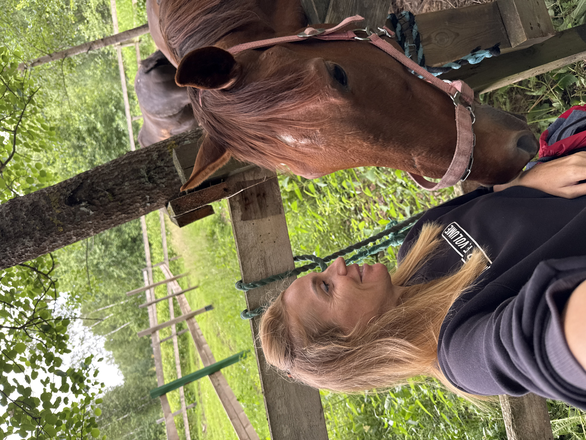 A woman with blonde hair pulled back is seated, smiling and looking to her left, towards the camera. She is wearing a black sweatshirt with white text on the chest that appears to read "F. VOLUME". In the foreground and to her right, the head of a brown horse with a pink halter is visible, facing downwards. Behind the woman and the horse, a wooden fence encloses a grassy area with trees and other vegetation. In the background, another dark-colored animal, possibly a horse or a donkey, is visible behind the fence. The scene appears to be outdoors, possibly a stable or a farm.