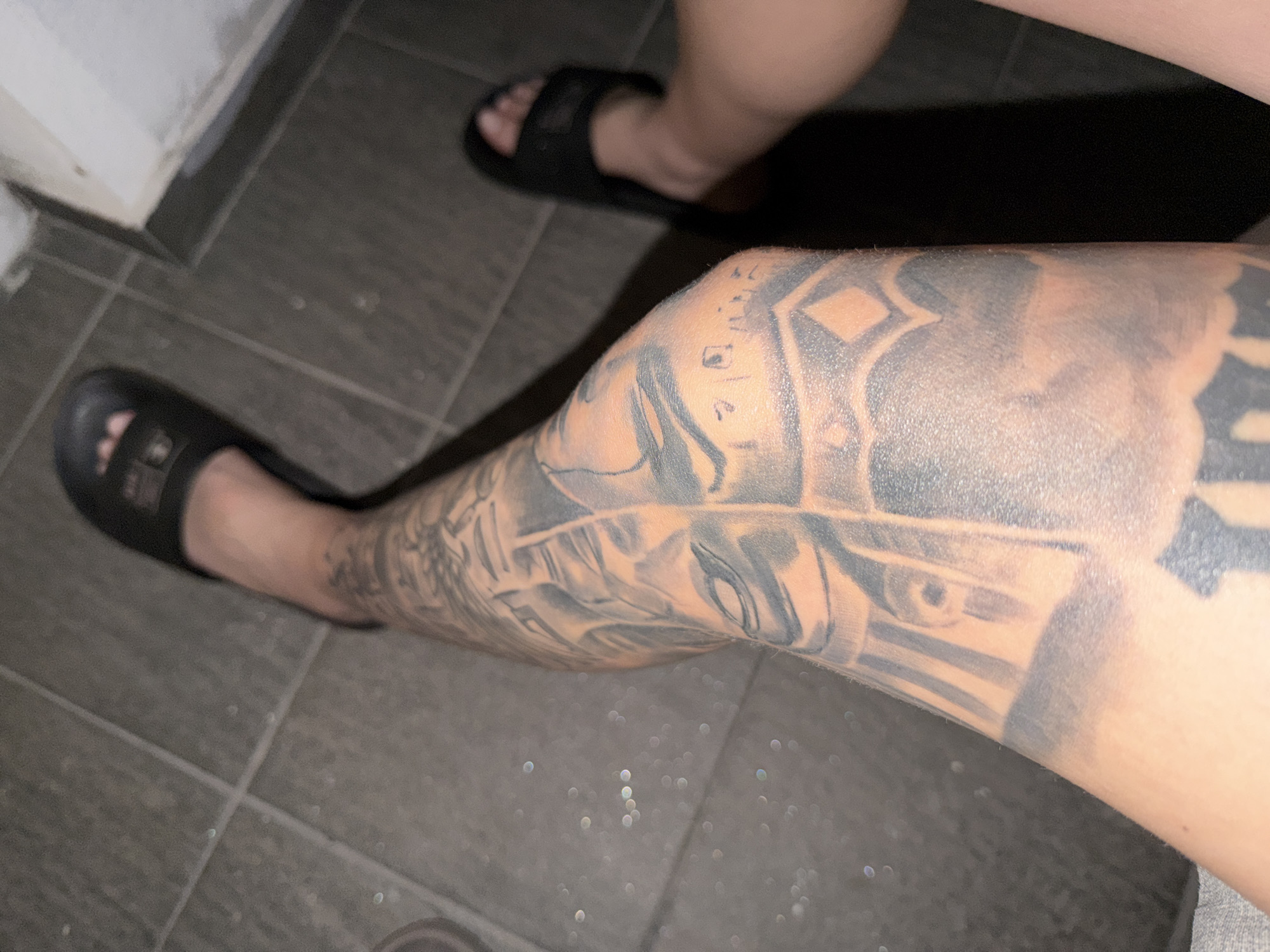 A downward-facing perspective captures the lower legs and feet of an individual on a tiled floor in the city of Plovdiv, Bulgaria. The floor is composed of grey rectangular tiles with visible grout lines. Dim lighting creates visible shadows beneath the feet. The subject's right leg (viewer's left) is adorned with an extensive black and grey tattoo sleeve from above the knee to the ankle, featuring an eye motif, geometric patterns, and abstract elements. The left leg (viewer's right) is less prominent, showing primarily the foot and lower calf. Both feet are clad in plain black slide sandals; a subtle detail is visible on the strap of the right sandal. A partial view of a white vertical surface, possibly a wall, is present in the upper right quadrant, meeting the tiled floor.