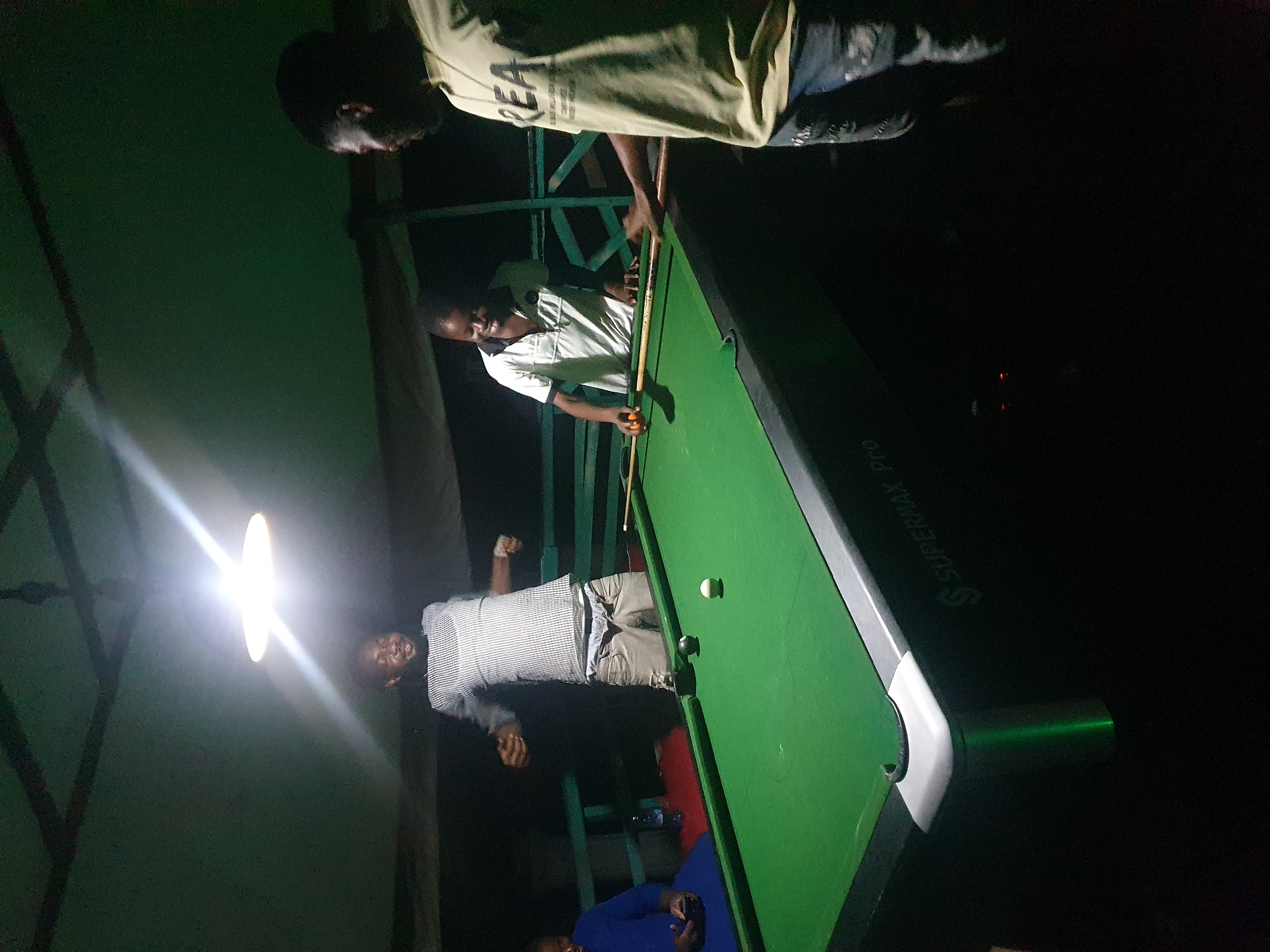 In Lokoja, Nigeria, at night, four men are gathered around a green pool table in an outdoor setting. The scene is illuminated by a bright overhead light, casting harsh shadows. Two men are actively engaged in a game of pool. One, wearing a striped shirt and khaki shorts, stands with his arms outstretched, appearing to be mid-celebration or reacting to a shot. The other, in a white polo shirt, is focused on the table, holding a pool cue. A third man, in a light-colored t-shirt with "REA" printed on it, stands to the right of the table, his hand extended as if ready to take his turn. A fourth individual, wearing a blue garment, is seated on a bench to the left, holding a phone. The pool table has a white and black ball on its surface. The surrounding environment suggests a casual establishment, possibly a bar or a recreational area, with wooden structures and railings visible in the background. The overall atmosphere is one of leisure and friendly competition.