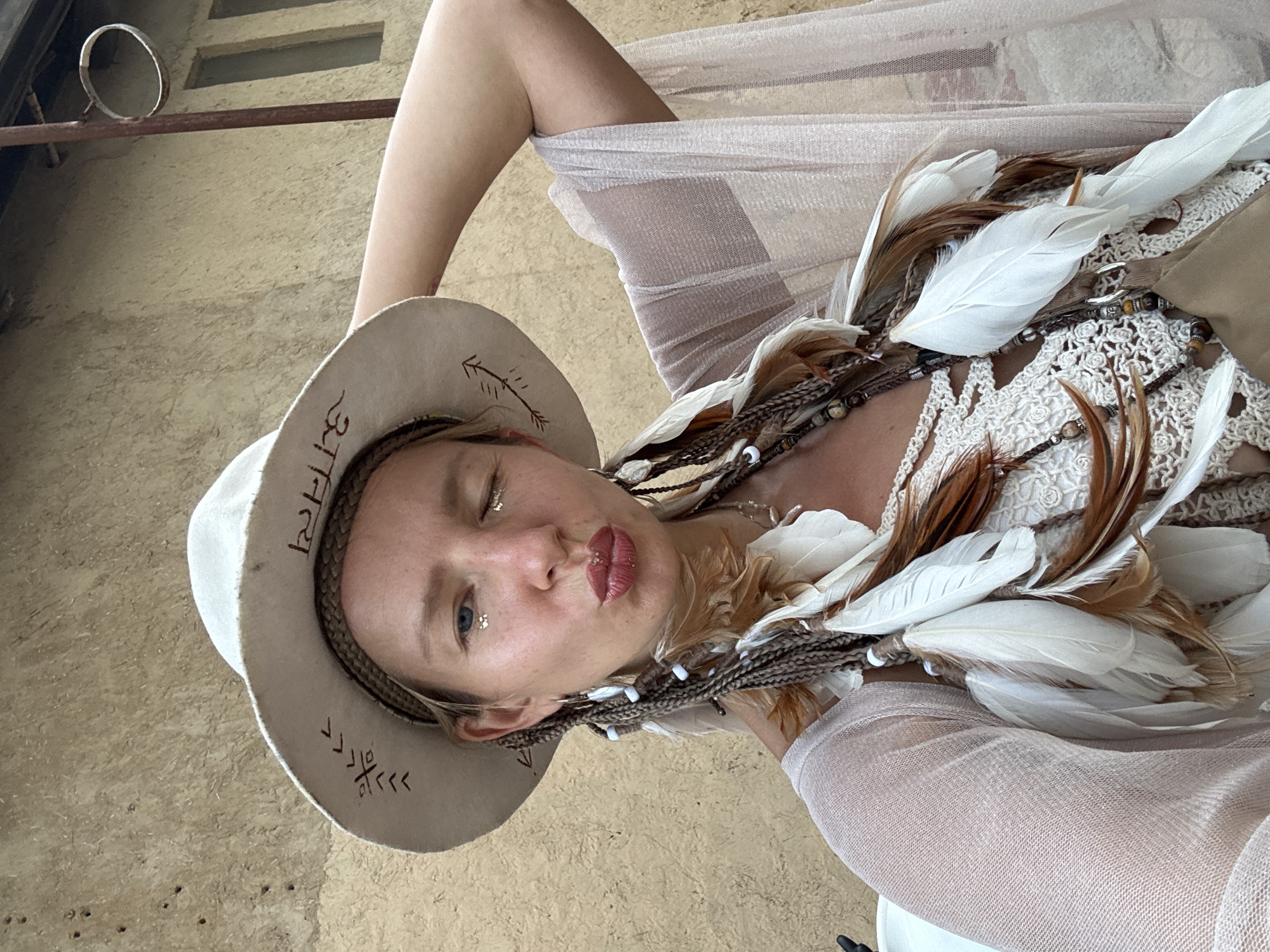 The image shows a woman with blonde hair styled in braids with feathers and beads, wearing a white wide-brimmed hat with embroidered symbols, and a crochet top. She has glitter on her eyelids and lips and is puckering her lips. She is also wearing a sheer, light-colored shawl over her shoulders. Her right arm is raised and bent, with her hand resting on the back of her head. The background is a textured, light-colored wall, likely made of stucco or adobe. The lighting suggests it is daytime. The setting appears to be outdoors. This is a portrait photograph taken by the subject of herself. There is no visual information in the image that places the scene in Mũi Né, Vietnam.