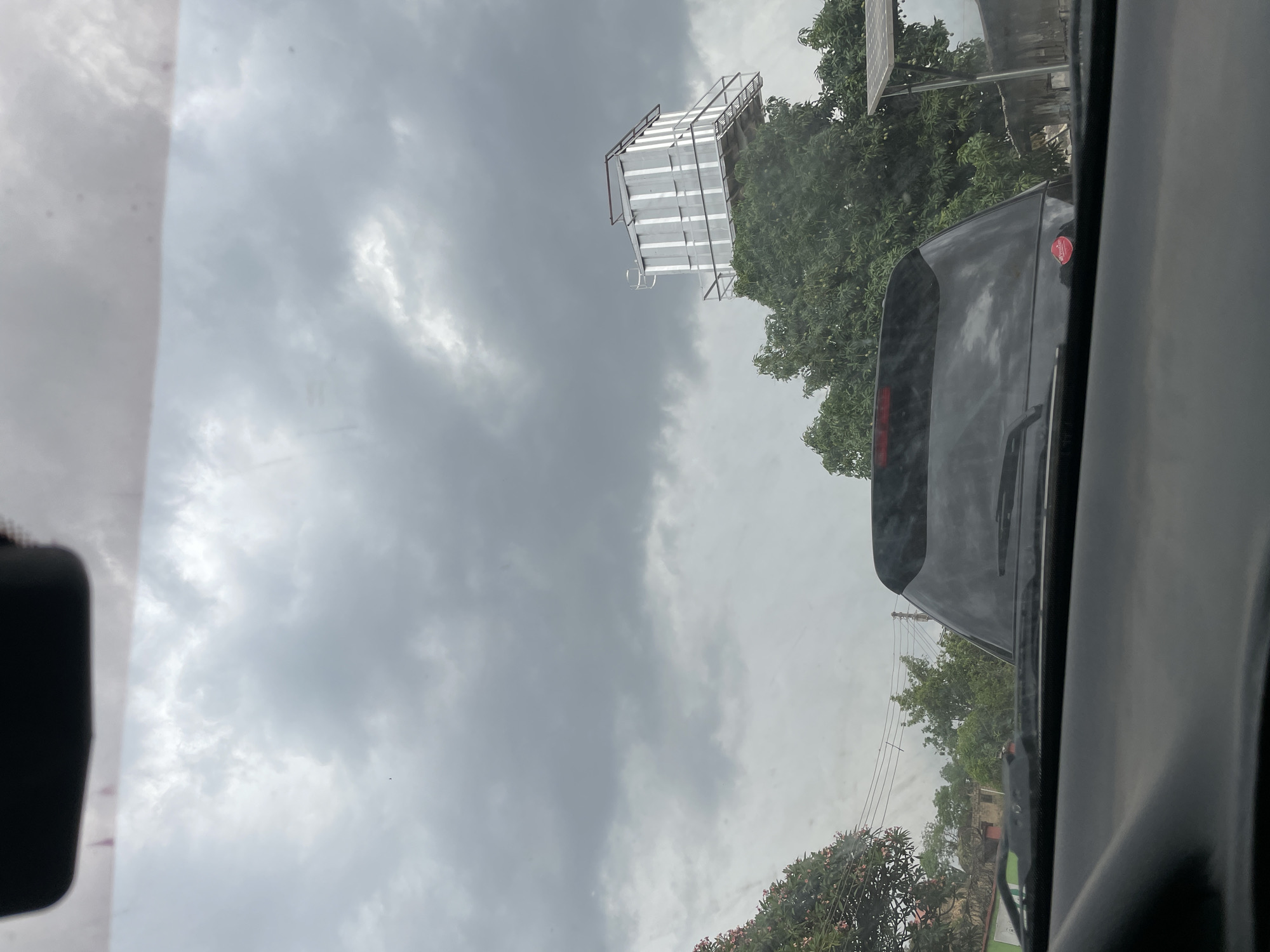 From inside a vehicle, the image shows a gray sky above Jos, Nigeria. A multi-tiered metal structure stands next to trees in the middle distance. In the foreground, part of another vehicle is visible.
