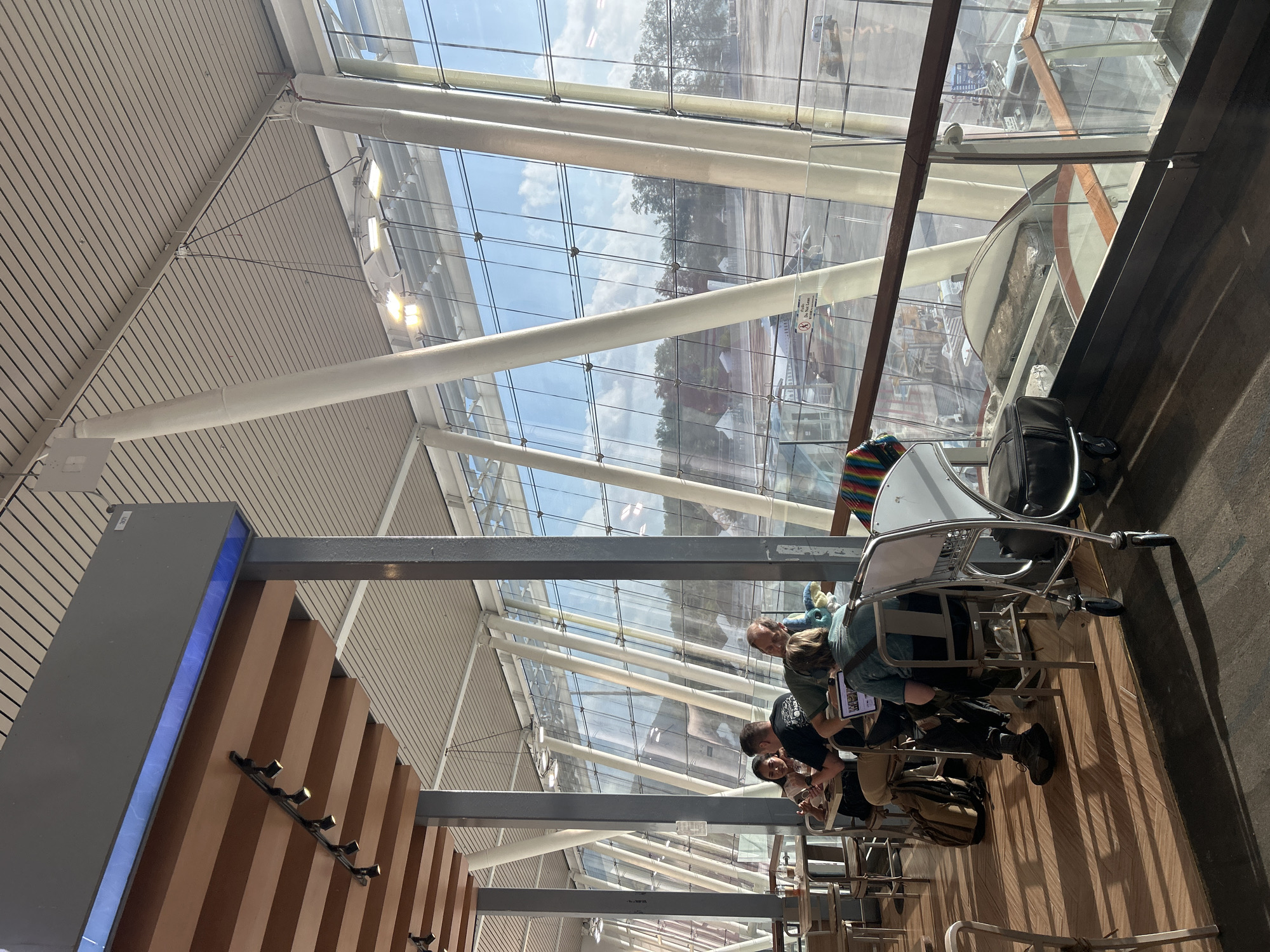 The image depicts an interior view of an airport terminal, likely in Tambon Mai Khao, Thailand, looking out through a large, slanted glass window. Several individuals are seated at tables, appearing to be patrons of a cafe or waiting area. One group of four people are engaged in conversation, with food and drinks on their table. Another person is seated with a laptop. The environment includes modern architectural elements such as a slatted ceiling, exposed beams, and prominent white support columns. The window offers a view of an airport tarmac, with a small jet plane visible in the distance, along with other airport infrastructure. The lighting suggests it is daytime, with sunlight casting shadows across the wooden floor.