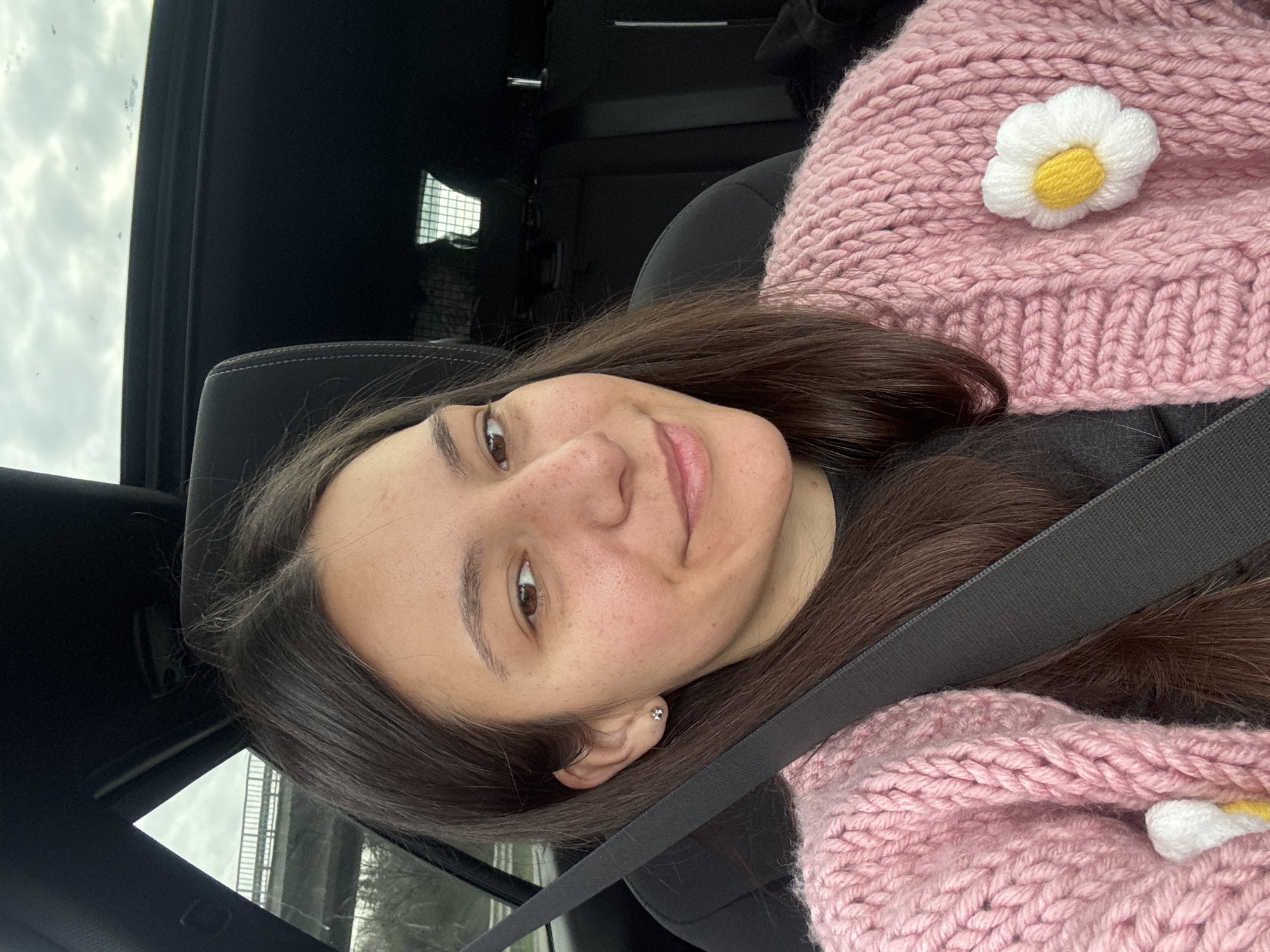 A young woman is seated inside a vehicle, likely a car, wearing a pink, chunky-knit scarf with a white and yellow flower embellishment. She has dark hair and is wearing a seatbelt. She is looking directly at the camera with a slight smile. The vehicle's interior is visible, including the roof and seat. The background seen through the windows is indistinct, but appears to be outdoors with cloudy skies. There is no indication of Kipra, Bulgaria, within the visual content of the image.