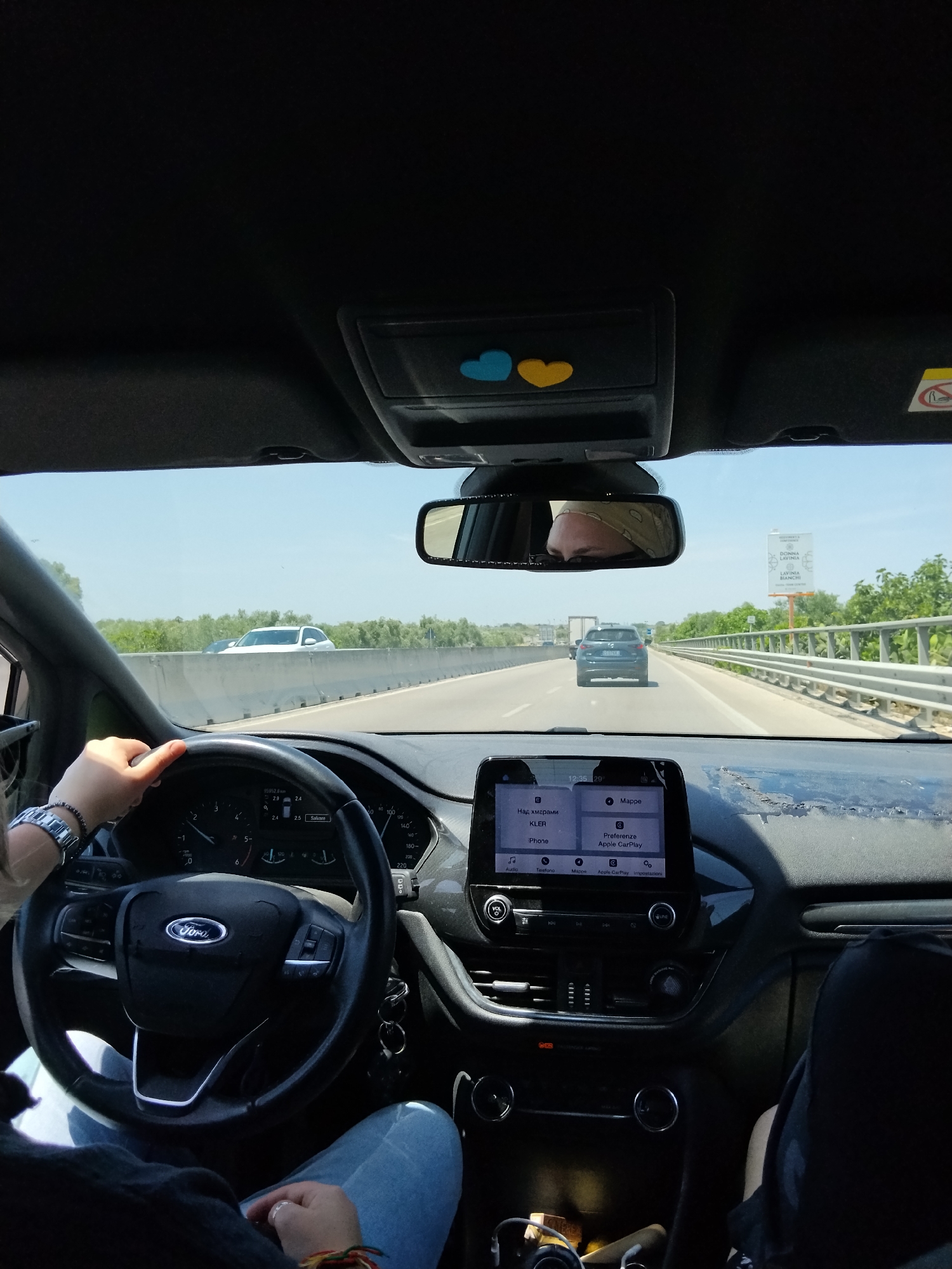 The image depicts the interior of a Ford vehicle from the front passenger seat, looking forward through the windshield onto a highway, specifically identified as being near Molfetta, Italy.
In the foreground, the steering wheel, bearing a Ford emblem, is grasped by two hands, presumably belonging to the driver. The driver's left wrist displays a silver-toned watch and a dark beaded bracelet. Behind the steering wheel, the instrument cluster shows a digital odometer reading "155952.8 km" and other indicators, including what appears to be a speedometer and a fuel gauge. Below the instrument cluster, a button labeled "Salvare" (Italian for "Save") is visible.
To the right of the steering wheel, the central infotainment screen displays a menu for Apple CarPlay with options in Italian and Cyrillic script, including "Audio," "Mappe," "Telefono," "Apple CarPlay," "Impostazioni," "Над хмарами" (Ukrainian for "Above the clouds"), "KLERR," and "iPhone." The time "12:35:29" is displayed at the top of this screen. The top surface of the dashboard exhibits significant wear, with material peeling and appearing damaged.
Below the infotainment screen, the climate controls and other buttons are visible. The lower portion of the image shows the passenger's lap, clad in blue jeans, with a hand wearing a ring on the left ring finger and a multicolored braided bracelet on the right wrist.
Above the windshield, an overhead console is visible, adorned with two heart-shaped stickers: one blue and one yellow. The rearview mirror reflects the driver's face, partially obscured by a light-colored, patterned head covering or bandana.
Through the windshield, a multi-lane highway is visible under a clear blue sky. Concrete barriers line the right side of the road, beyond which are lush green trees and bushes, possibly olive groves. Several vehicles are ahead on the highway, including a white SUV on the left lane and a dark blue sedan directly in front of the Ford. Further in the distance, a large truck is also visible. On the right side of the highway, a large billboard prominently displays "DONNA LAVINIA" and "LAVINIA BIANCHI," identifying the location as being near Molfetta, Italy.
